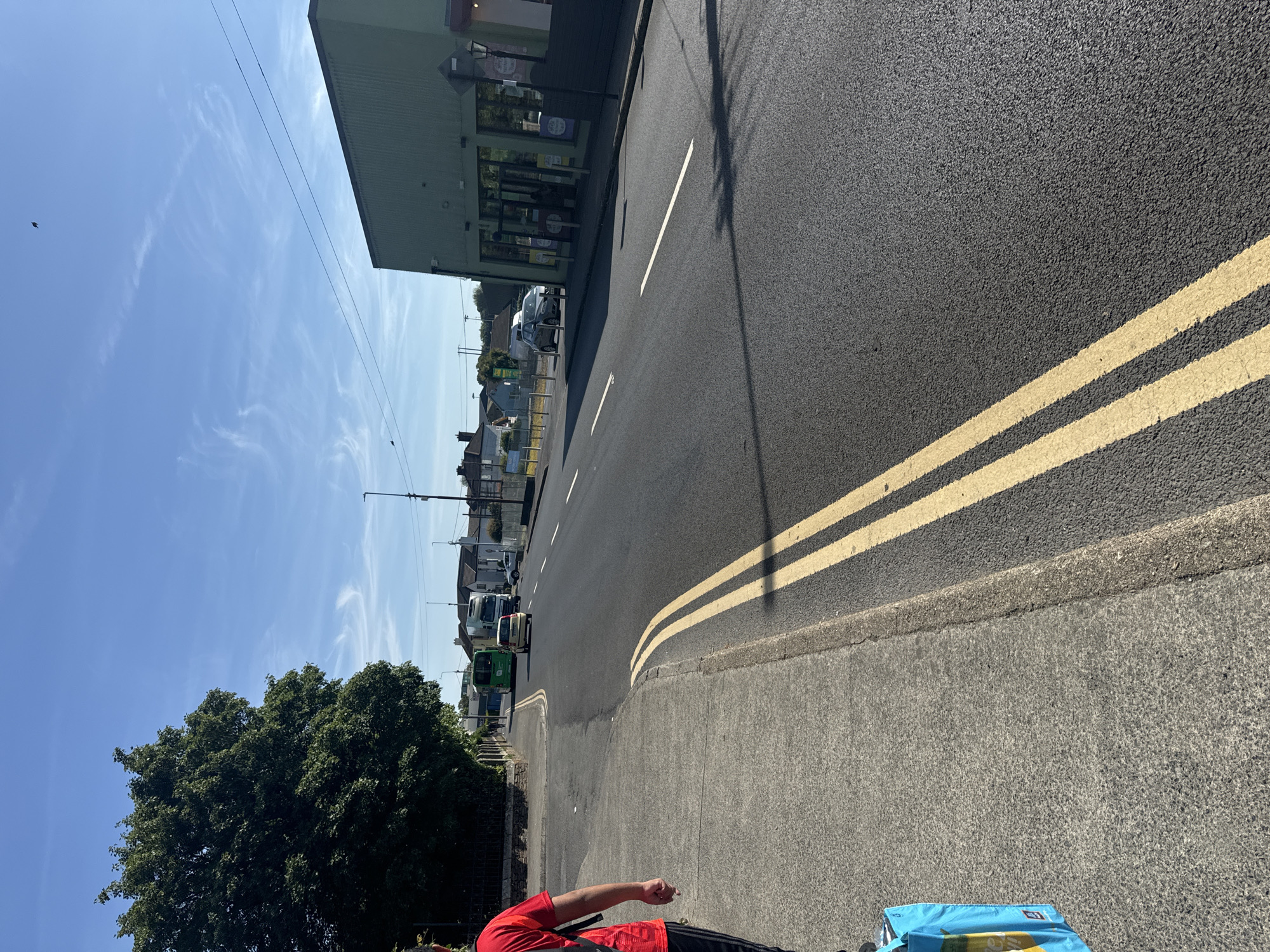The image shows a street scene in Waterford, Ireland. On the left side of the image, a person wearing a red t-shirt and black trousers is walking on a concrete sidewalk, carrying a blue shopping bag. Behind the person, a large green tree is visible. The street is paved with asphalt and has double yellow lines painted on it. Several vehicles, including a green bus and a white truck, are driving on the road. On the right side of the image, a green building with large windows and an entrance is situated along the street. The sky is clear and blue with some wispy clouds. The scene appears to be a typical day in a commercial or residential area.