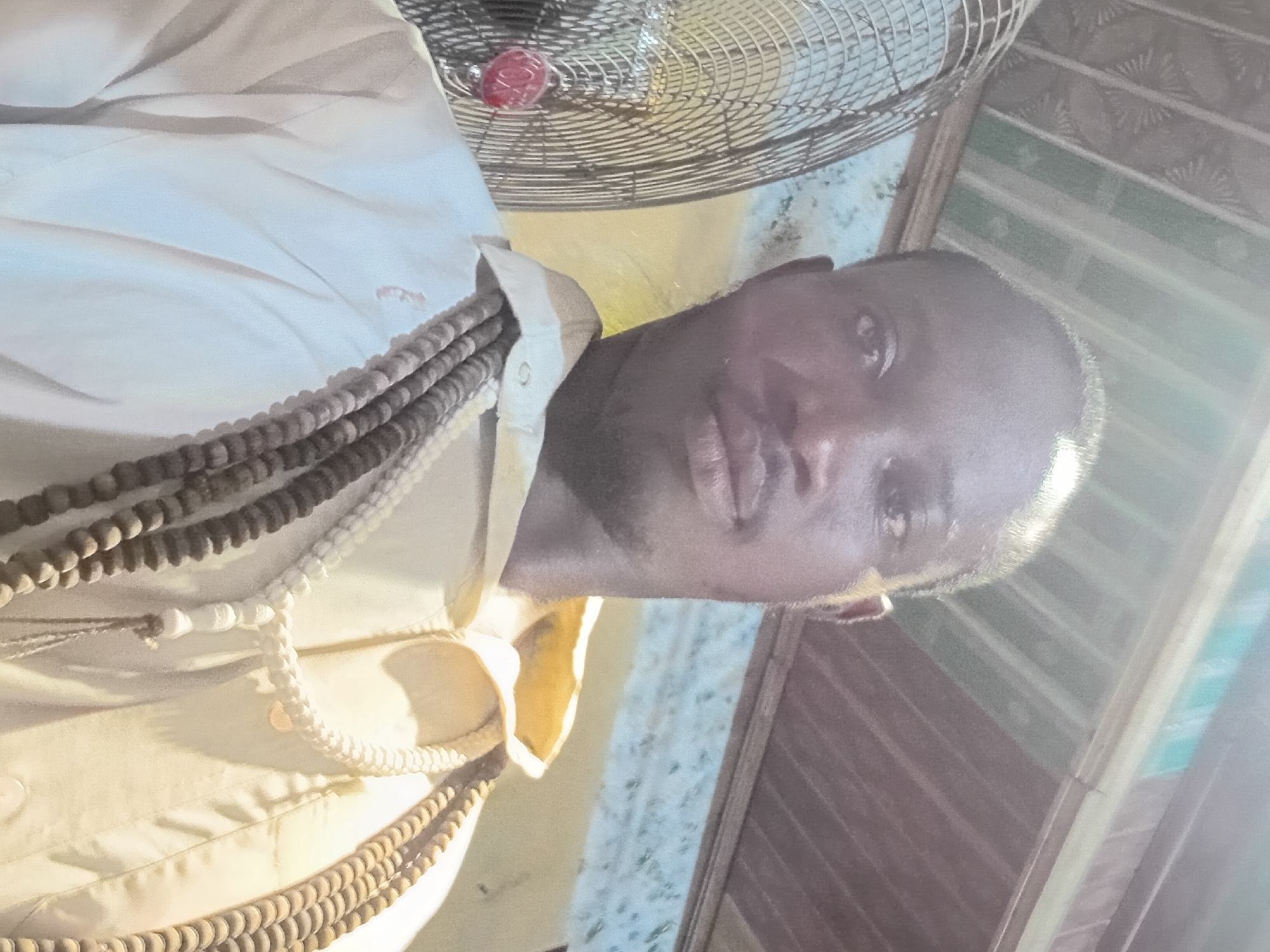 A selfie taken from a slightly upward angle shows a Black man from the chest up. He has dark skin and is wearing a light-colored collared shirt. Around his neck, he wears several strands of prayer beads, varying in size and color from light beige to dark brown. To the left of the man, a large, metallic fan with a red accent is visible. The background suggests an indoor setting, with a corrugated metal ceiling above and a patterned wallpaper or wall covering behind. The lighting is bright, casting some glare and shadows.