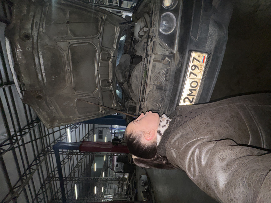 A woman in a brown jacket looks into the engine bay of a dirty car with its hood open. The car has a Russian license plate "2MO 797 RUS" visible. The scene appears to be inside a repair shop, with a metallic ceiling structure and a red lift in the background. The environment looks dimly lit.