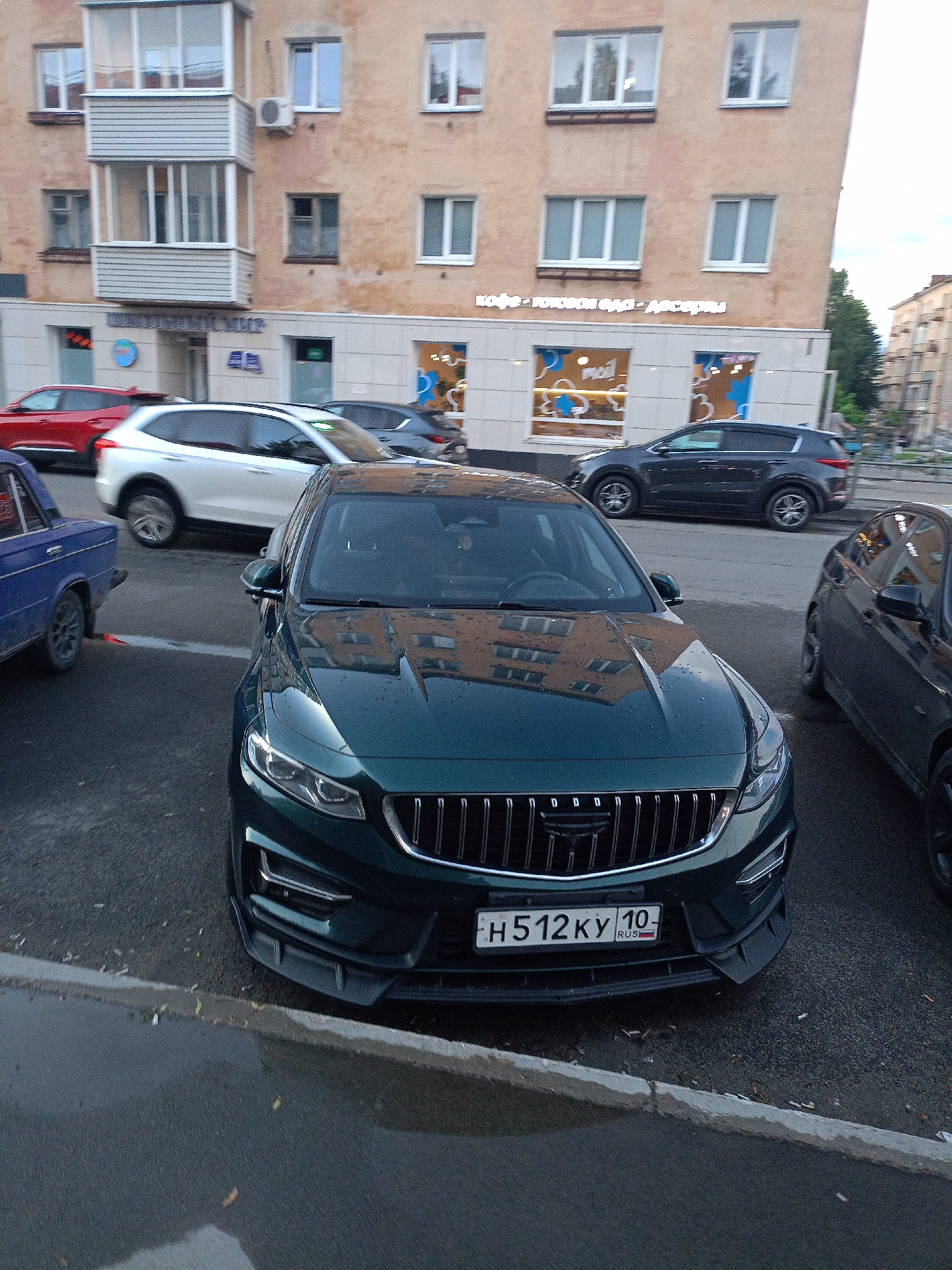 The image shows a street scene with several cars parked and driving on a wet asphalt road. In the foreground, a dark green car with a license plate that reads "H512KY 10 RUS" is prominently featured. The car has a distinctive grille and front bumper. Behind it, a blue car is partially visible. Further back, a white SUV and a dark gray SUV are also present. The background consists of a multi-story building with various windows and a balcony. Some signage is visible on the building, including what appears to be "КАФЕ" (cafe) and "ДЕСЕРТЫ" (desserts). The overall environment suggests an urban setting, possibly a street in front of a commercial establishment, with overcast weather given the wet conditions.