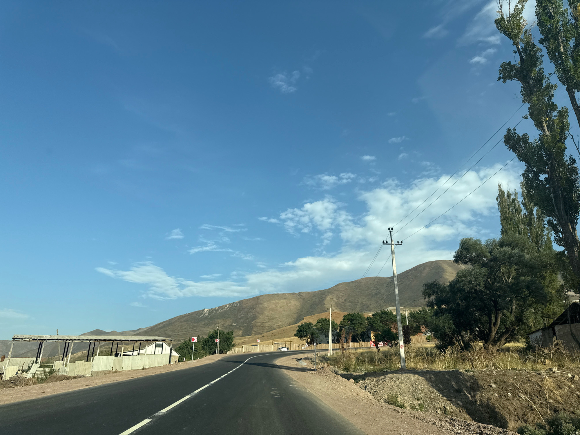 The image depicts a winding asphalt road passing through a rural, mountainous landscape. The road is bordered by dry, rocky terrain and sparse vegetation. On the left side of the road, there is a simple, open-sided structure with a corrugated metal roof, possibly a shelter or storage area. Further down the road, a white building with a dark roof is visible.
The right side of the road features a cluster of tall, leafy trees, some of which are quite large. A utility pole with power lines runs along the roadside. In the background, rolling, dry, brown hills dominate the horizon under a clear blue sky with scattered white clouds. The overall impression is one of a remote, somewhat arid, and undeveloped region. There are no people or animals visible in the image. The image is taken from the perspective of someone in a vehicle moving along the road.