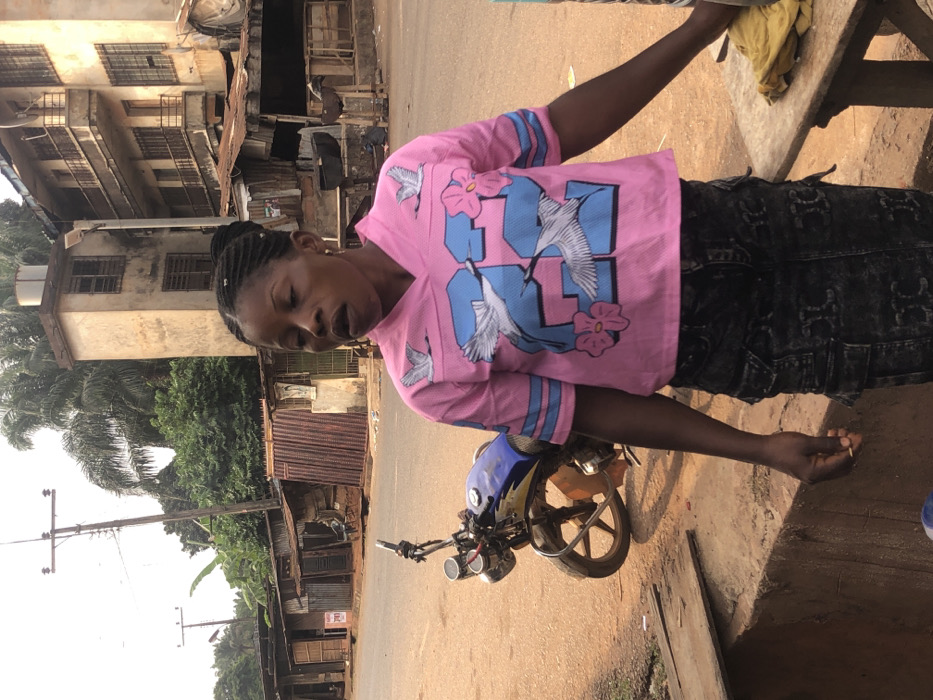 This image depicts a street scene in Neni, Nigeria. A dark-skinned female, with braided hair pulled into an updo, stands in the foreground, slightly right of center. She wears a pink short-sleeved V-neck shirt adorned with blue stripes on the sleeves, a large blue "32" graphic, and white crane and pink flower designs. She is also wearing a dark, distressed denim cargo skirt. Her mouth is slightly open, and her gaze is directed downwards and to the right of the frame. Her left hand loosely holds a small, light-colored, cylindrical object. To her immediate right, a portion of a wooden surface, possibly a table or counter, is visible.
In the mid-ground to the left, a blue and black motorcycle is parked on a reddish-brown dirt shoulder adjacent to a paved road. The background features a blend of structures and vegetation. On the left, a series of single-story buildings, characterized by corrugated metal roofs and varied wall materials, line the street. On the right, a multi-story concrete building, appearing weathered, stands prominently, alongside a taller, narrower concrete structure. Dense green trees and palm foliage are visible behind these buildings. Several utility poles with overhead wires are present against the bright sky. The immediate foreground consists of reddish-brown dirt, with a low concrete barrier or drainage ditch on the left side.