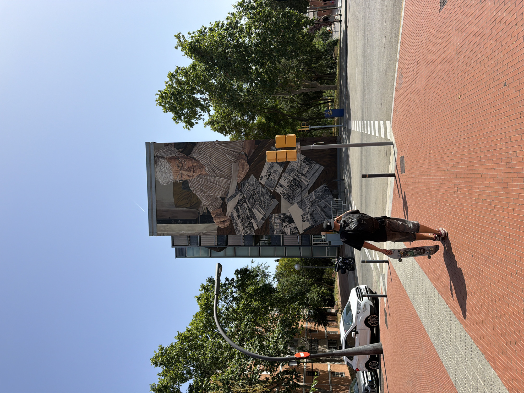 A wide-angle outdoor shot captures a sunlit urban scene in Barcelona, Spain, dominated by a building adorned with a large mural. The mural depicts an elderly woman with gray hair and glasses, seated and looking at photographs spread on a table. Below her, a collage of black and white images shows scenes of people.
In the foreground, a person wearing a black t-shirt, shorts, a backpack, and a baseball cap walks away from the camera, carrying a skateboard. They are on a red brick sidewalk, which is intersected by a grey and white striped pedestrian crossing in the midground. To the left of the sidewalk, a white SUV is parked, and a street lamp with a red circular sign attached to it stands tall.
The background reveals more trees and buildings, with a clear blue sky overhead. Traffic lights, one yellow and stacked, are visible on a pole near the pedestrian crossing. The scene suggests a typical day in a city with a blend of residential buildings, public art, and pedestrian activity.