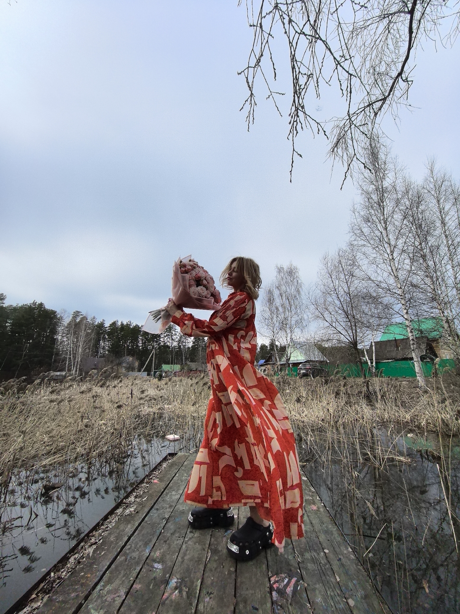 A woman in a long, flowing red and tan patterned dress stands on a wooden dock, holding a bouquet of pink flowers wrapped in pink paper. She is wearing black shoes with decorative embellishments. The dock appears to be situated on a body of water surrounded by tall, dry grasses and reeds. In the background, there are scattered trees, some with bare branches, and a few small buildings, including one with a distinctive green roof. The sky is overcast, and bare branches are visible in the upper right corner of the frame. The photo was taken in the city of Noviy, Russia.