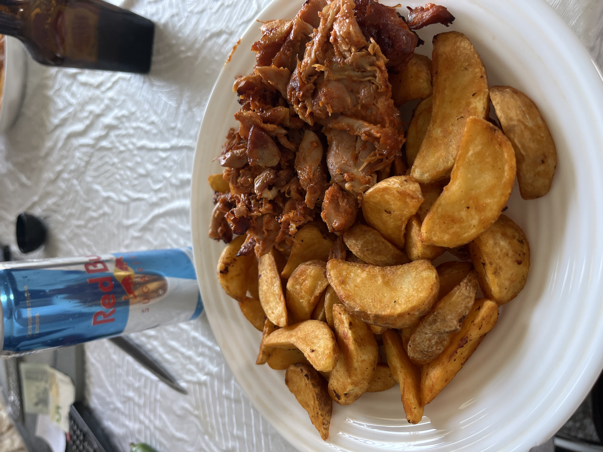 This close-up, top-down image showcases a casual meal laid out on a white, textured tablecloth, likely indoors at a dining table.
The central subject is a large white plate abundant with food. On the left side of the plate, there's a generous portion of shredded, well-cooked meat, possibly chicken or pork, coated in a reddish, savory sauce. Some pieces appear caramelized or slightly charred, suggesting a flavorful preparation similar to gyros, shawarma, or pulled meat. Complementing the meat, the right side of the plate is filled with numerous golden-brown potato wedges, some showing visible seasoning or a crispy texture.
Adjacent to the plate on the left, a blue and silver Red Bull energy drink can is prominently visible. The text "Red Bull" is clearly legible in its distinct font, along with "SUGARFREE" and smaller Cyrillic text, likely providing product details. Further to the bottom-left, partially obscured by the can, is a 5 Bulgarian leva banknote, identifiable by the number "5" and a portion of the Bulgarian lion emblem. This detail serves as a strong location cue, aligning with the provided context of Zlatograd, Bulgaria. Additional background elements include a dark, partially visible object at the top-left, and a black gridded object, possibly a keyboard, along with a small green item at the very bottom-left.
The scene depicts a meal prepared and ready for consumption, suggesting a lunch or dinner setting. No people are visible, so no specific activity or emotions are directly portrayed. The overall impression is one of a hearty and satisfying plate of food, typical of a casual dining experience.