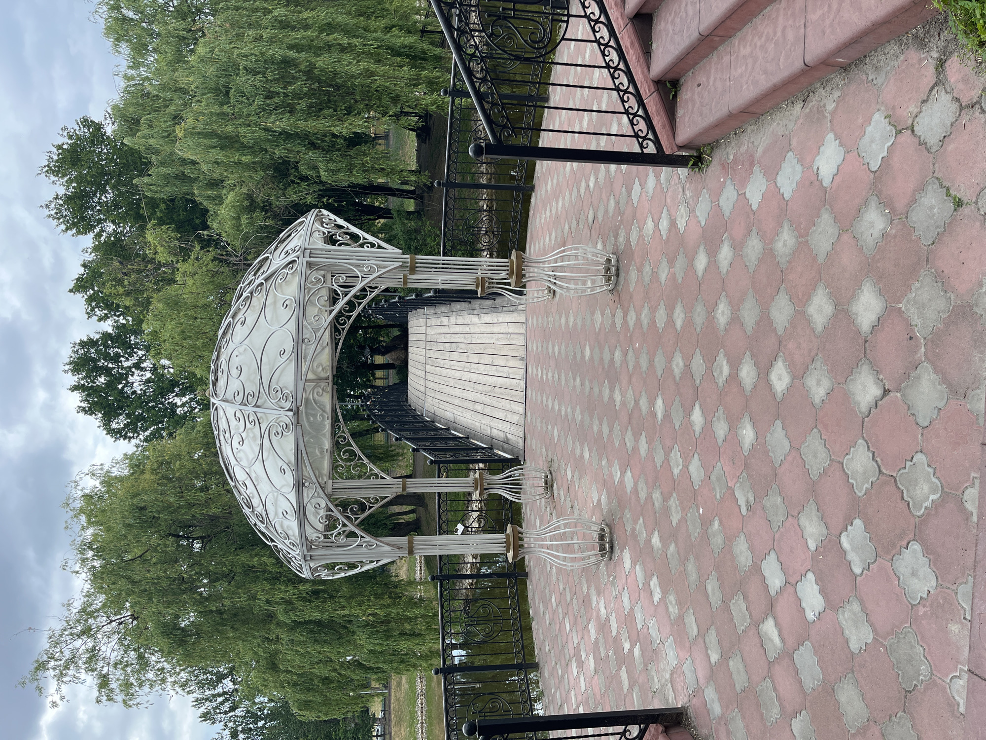 The image shows an ornate white gazebo with a domed roof situated on a paved walkway. The walkway is made of red and gray interlocking pavers in a quatrefoil pattern. In front of the gazebo, four decorative white metal structures resembling vases stand on the pavement. A wooden bridge extends from the walkway, crossing over a dark body of water. The bridge is bordered by a black metal railing with decorative scrollwork. Beyond the water, a dense line of green trees, including weeping willows, forms a backdrop. The sky above is overcast with gray clouds. To the right of the gazebo, a set of red-brown stairs with black metal railings leads up to the walkway.