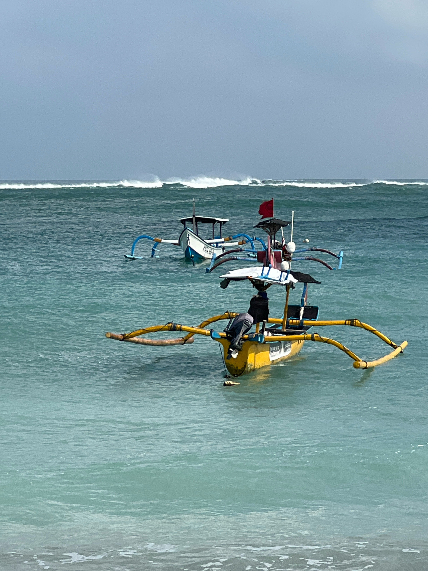 This image shows two traditional Balinese fishing boats, known as jukungs, on the turquoise waters off Kuta Beach in Bali, Indonesia. The boat in the foreground is yellow and features an outrigger system with prominent yellow bamboo outriggers. A person wearing a dark jacket and gray pants is seated on the edge of this boat, facing away from the camera and towards the water. The boat behind it is white with blue accents and also has outriggers. In the background, large waves are breaking, indicated by white foam crests against a hazy, overcast sky. A red flag is visible on the rear of the yellow boat, possibly indicating a warning or a specific fishing area. The water is calm around the boats, with small waves lapping at the shore in the lower portion of the frame.