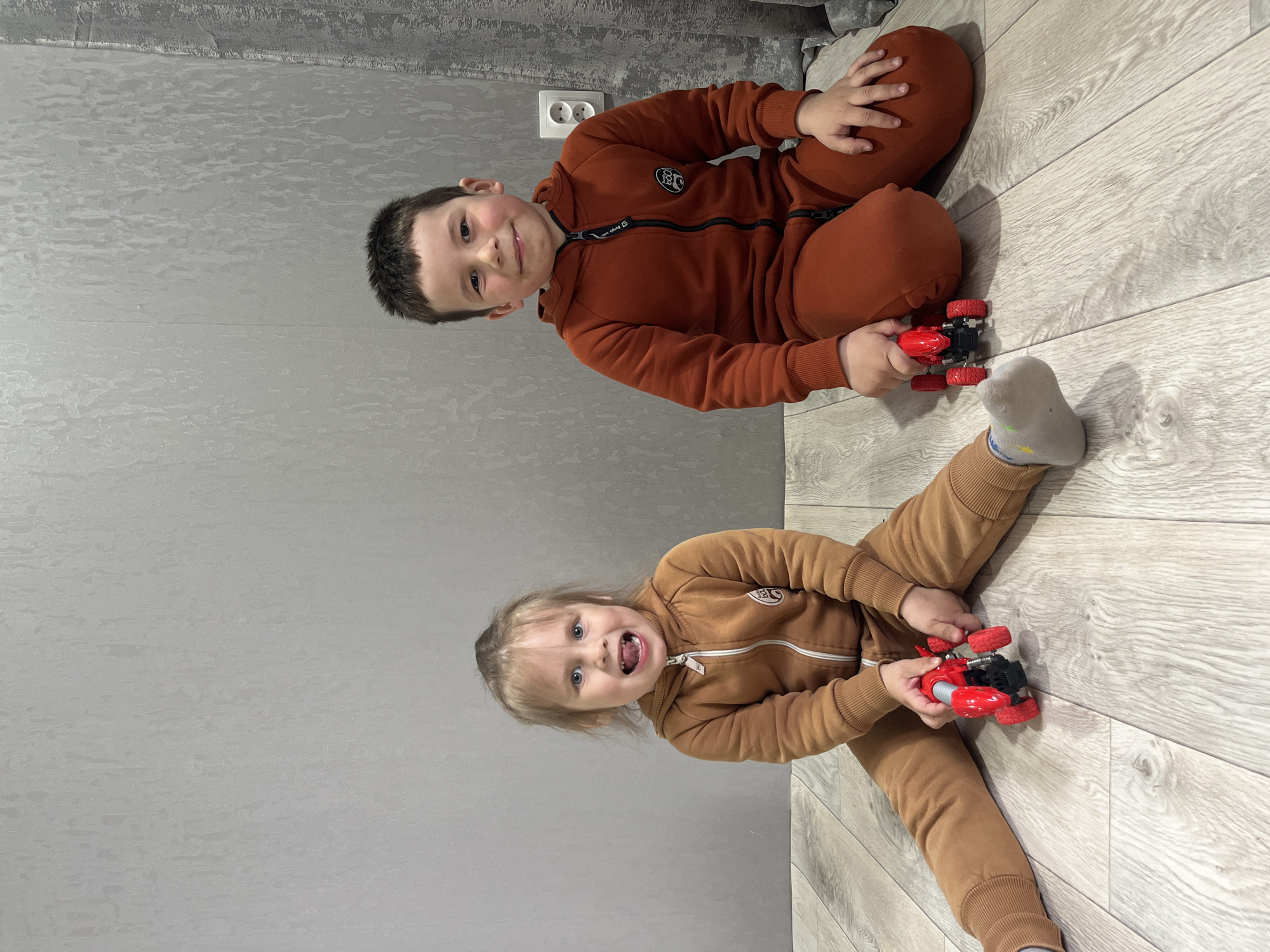 Two young children are captured in an indoor setting in Chaikovsky, Russia, sitting on a light-colored wooden floor next to a textured grey wall. The image appears to be rotated 90 degrees counter-clockwise.
A boy, positioned on the right (when oriented correctly), wears a rust-colored tracksuit. He has short, dark hair and gazes towards the camera with a gentle smile. His tracksuit features a black zipper and a circular patch on the chest with white text, clearly visible as "BOOM" and "ORIGINAL". He is holding a red and black toy car in his hands, which are resting on his crossed legs.
To his left, a girl in a light brown tracksuit sits with her legs spread out. She has blonde hair pulled back and looks directly at the viewer with a wide-open mouth, revealing a playful, toothy grin. She also holds a red and black toy car, identical or very similar to the boy's. Her right foot, wearing a grey sock, is visible.
Both children appear happy and engaged, seemingly pausing their play with the toy cars to pose for the photograph. An electrical outlet with two circular sockets, common in European and Russian settings, is visible on the grey wall behind the boy. The lighting suggests it's daytime.