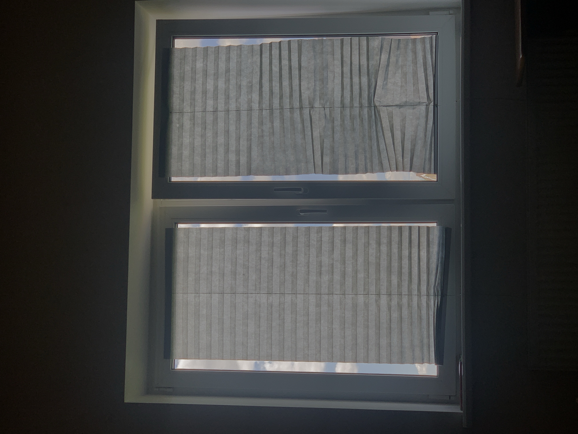 The image shows a closed window with two pleated blinds. The blinds are gray and appear to be made of fabric. The window is divided into two panes, and each pane has a blind. The blinds are slightly crumpled, especially on the right side, suggesting they may be old or damaged. Through the blinds, a blue sky with white clouds is visible. The overall lighting suggests it is daytime, possibly late afternoon or early morning due to the shadows. The scene is indoors, and the dark area at the bottom of the image may be a windowsill or furniture. No people are visible. There is no information within the image to suggest it is located in Minsk, Belarus.