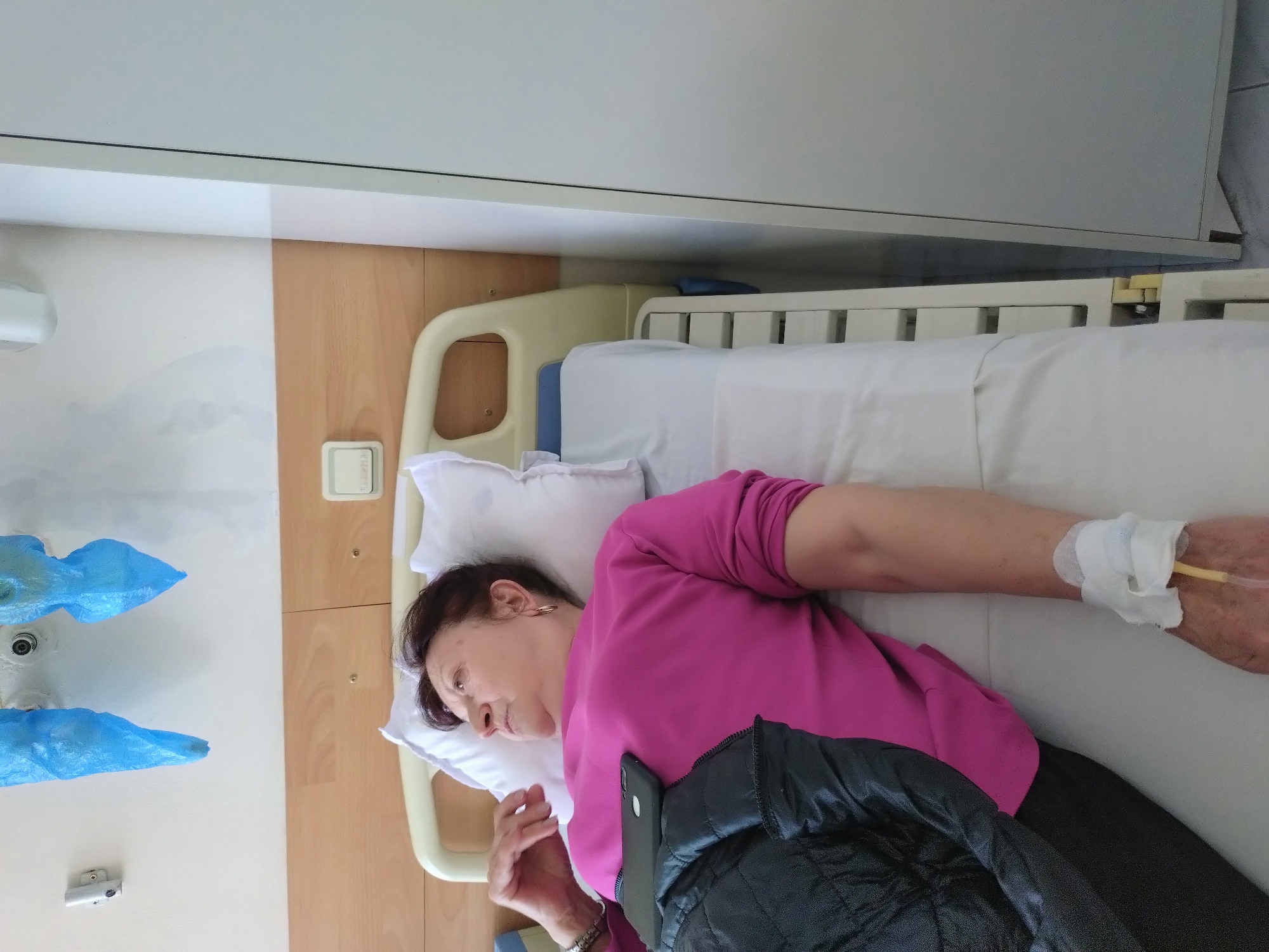 An elderly woman lies in a hospital bed, wearing a pink shirt. An IV line is connected to her right arm. The bed is equipped with rails. Her left hand is visible, and a dark jacket lies next to her. A smartphone is near her jacket. The room has light-colored walls and wooden paneling. Blue shoe covers are visible near a sink. The location is likely a hospital room in Stara Zagora, Bulgaria, based on the assumption that the user has knowledge of the city.