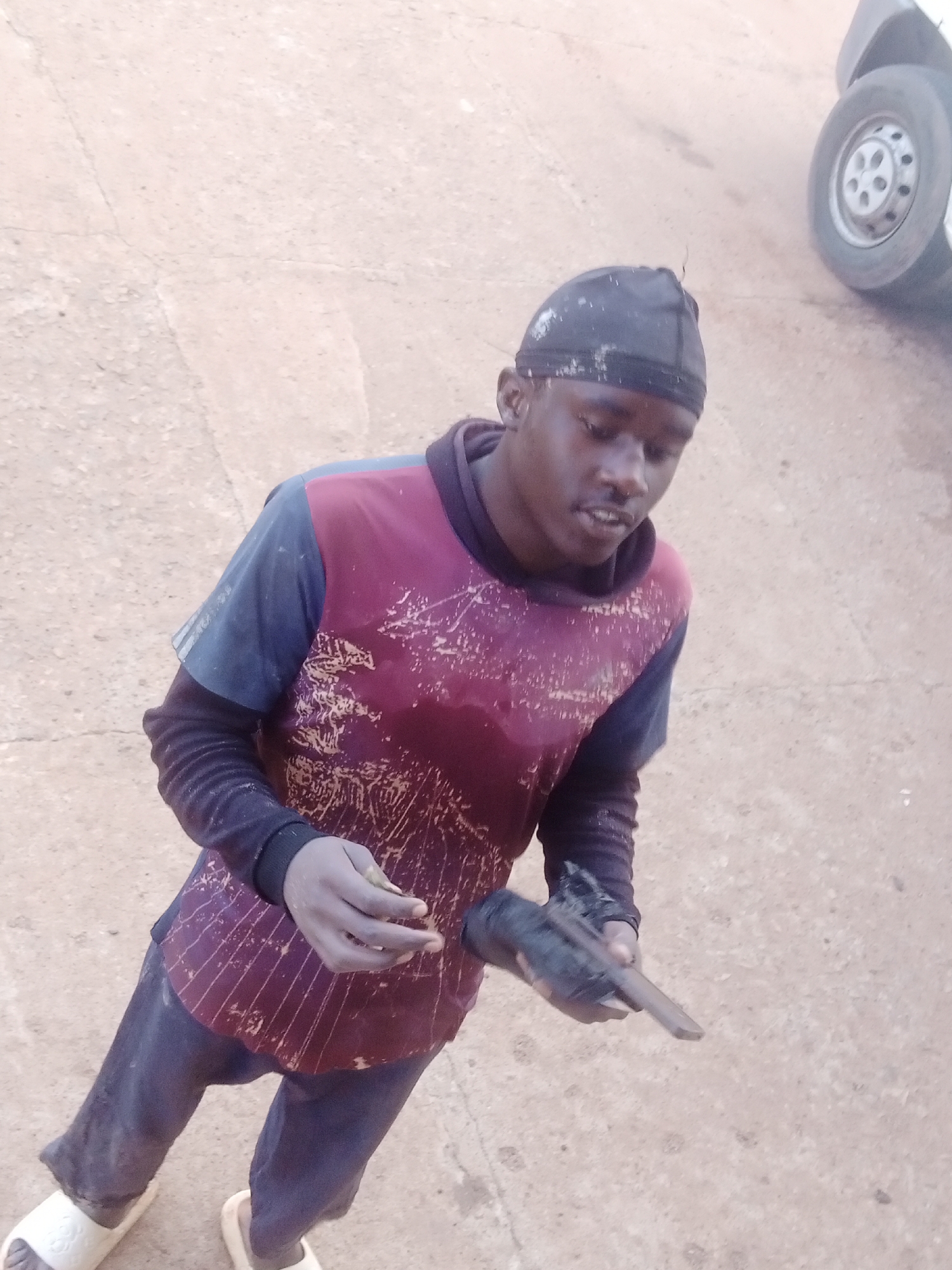 A young Black male wearing a black durag, a maroon and blue long-sleeved shirt, and dark pants stands on a dirt path. The male is holding a small, dark object in his hands. A white van is visible in the background, and the ground is a light brown dirt with some cracks. The scene appears to be outdoors in an urban environment.