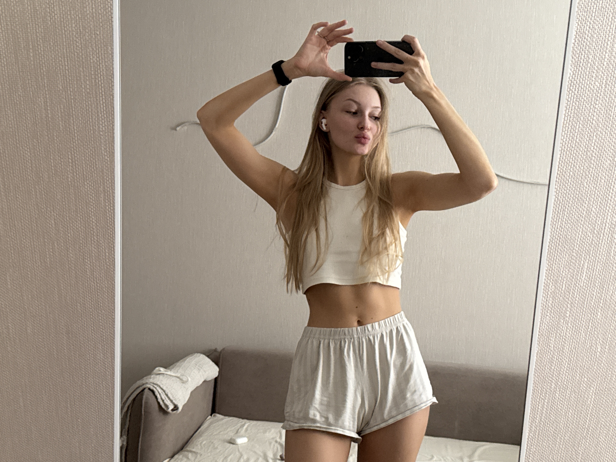 In an indoor setting, a young woman with blonde hair and fair skin stands in front of a mirror, taking a selfie with her smartphone. She is wearing a white crop top and light-colored shorts. She has white wireless earbuds in her ears and a black watch on her left wrist. Her arms are raised, holding the phone above her head, and she is puckering her lips. The background shows a textured wall and a glimpse of a sofa with a blanket. This scene is not in Perm, Russia.