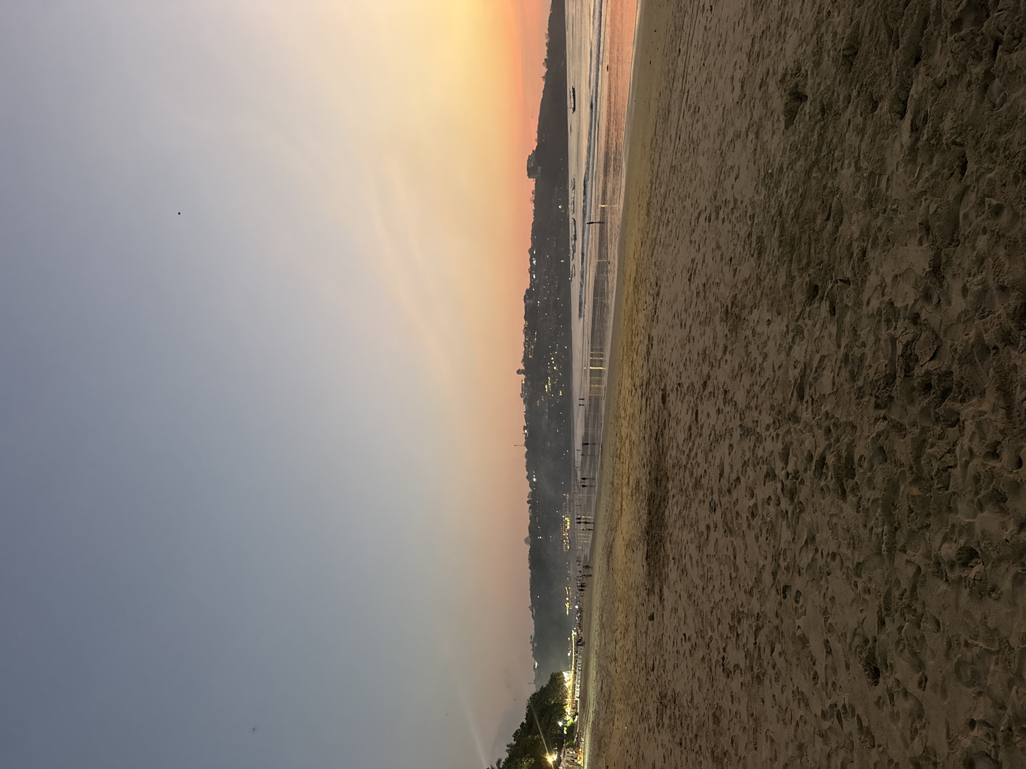An eye-level, long shot captures a twilight scene at Kuta Beach in Kuta Selatan, Indonesia. The sky transitions from a pale blue at the top to a warm orange and pink gradient near the horizon, indicating the setting sun. A few specks, possibly birds or insects, are visible against the sky.
The foreground is dominated by the sandy texture of the beach, showing numerous footprints. In the midground, the calm ocean reflects the colors of the sky. Small waves gently lap the shore. Several figures, mostly appearing as silhouettes due to the low light, are scattered along the water's edge and further up the beach. Some appear to be walking, while others may be standing.
Along the coastline in the background, a hilly landscape is dotted with numerous lights from buildings and structures, suggesting a populated area. Trees are visible on the left side of the frame, with some illuminated lights among them, possibly from restaurants or stalls. Several boats, some appearing as dark shapes, are visible in the water. The overall atmosphere is serene and peaceful, typical of a beach at dusk.
