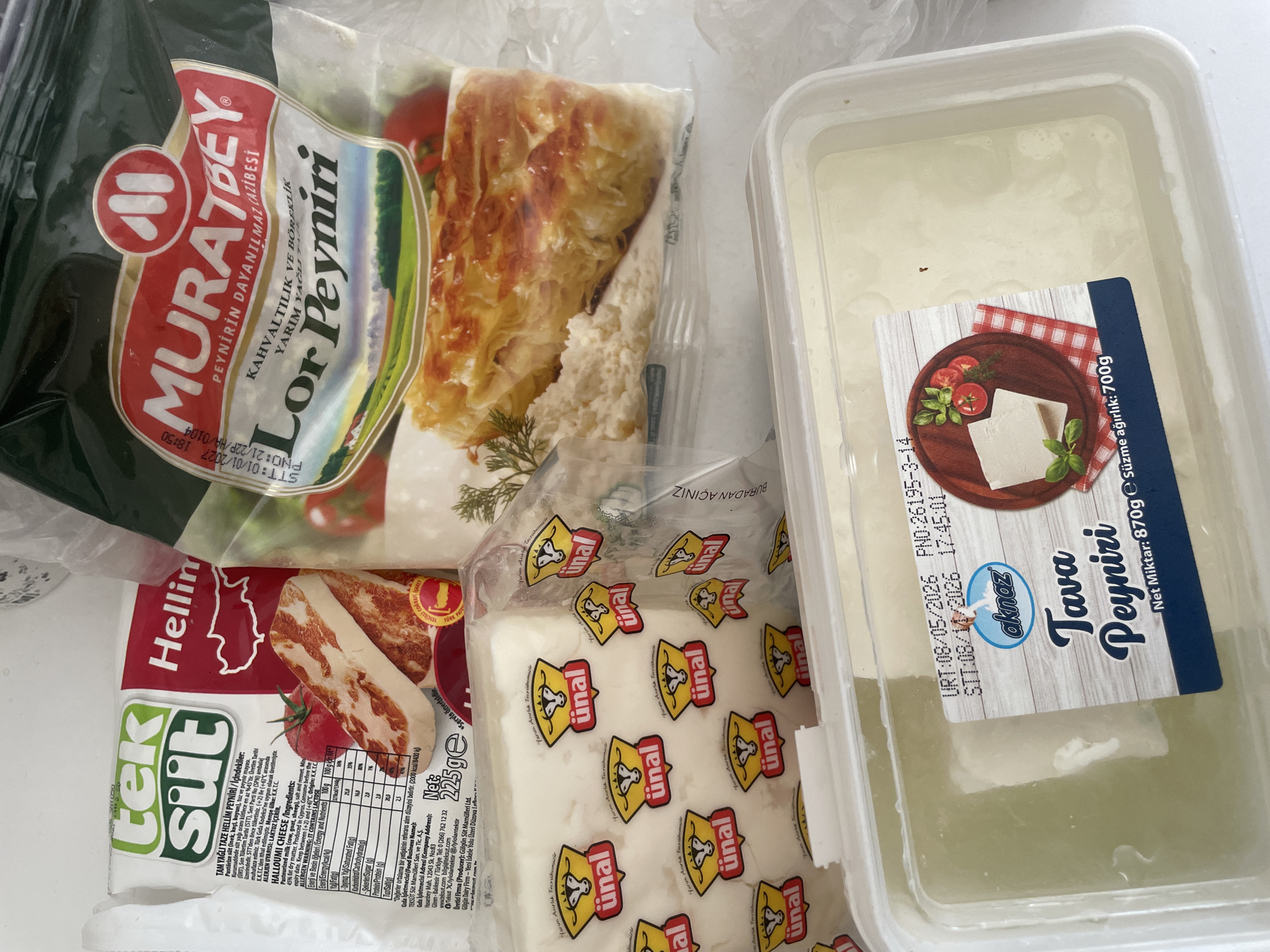 The image displays a collection of cheese products, likely purchased from a supermarket. The foreground features a white plastic container filled with a pale yellow, liquid substance, which appears to be brine or whey, and contains a block of cheese. A label on this container indicates "akmaz TAVA PEYNIRI" and provides weight information of "700g". In the middle ground, several individually wrapped cheese portions are visible, each bearing the "ünal" logo with a cow icon. Behind these, a package of "TEK SÜT Hellimi" cheese is prominent, showing nutritional information and an image of grilled halloumi. To the right, a package of "MURATBEY Lor Peyniri" with an image of pastry is also visible. The overall setting suggests a domestic environment, possibly a kitchen counter, with the products arranged for storage or use. The products are typical of Turkish dairy brands. The location of Başakşehir, Turkey, is not discernible from the image itself but is a known source for such products.