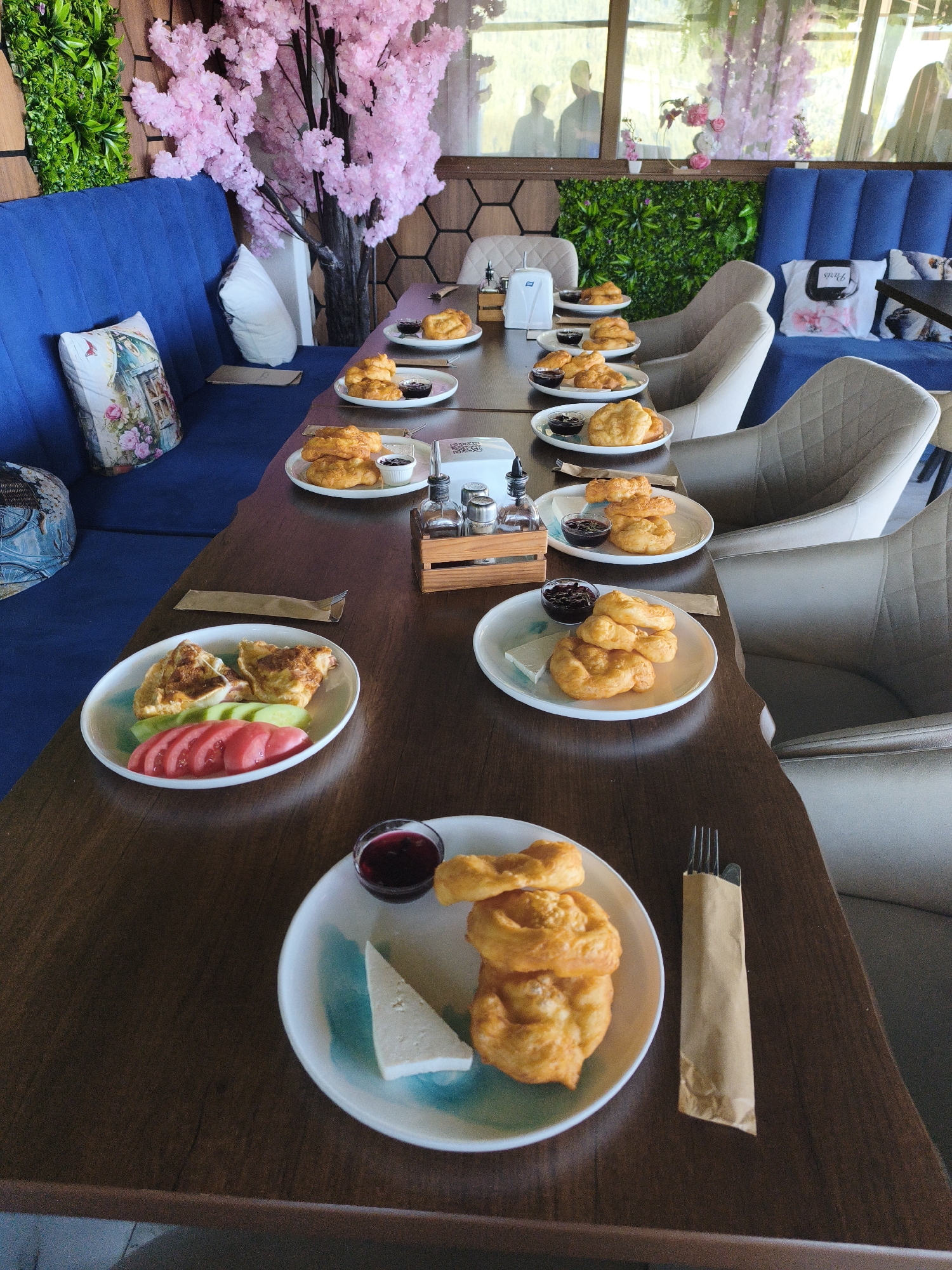 The image depicts a table setting in what appears to be a cafe or restaurant in Sarnitsa, Bulgaria. The table is made of dark wood and is laden with several plates of food, primarily pastries and some fresh vegetables. The pastries are golden brown and appear to be a type of fried dough, some are stacked and others are arranged in a circular pattern. Each plate with pastries also contains a small cup of dark jam and a piece of white cheese, except for one plate which features sliced tomatoes and cucumbers alongside some fried pastry. There are also salt and pepper shakers and a bottle of oil or vinegar on a wooden tray.

The seating area is characterized by comfortable-looking blue upholstered benches and light-colored chairs with textured fabric. In the background, there is a wall decorated with artificial green foliage and a prominent artificial tree with pink blossoms, suggesting an effort to create a visually appealing and perhaps themed ambiance. Through a window in the background, two silhouetted figures can be seen. The overall scene suggests a breakfast or brunch setting, with multiple servings prepared and awaiting consumption.