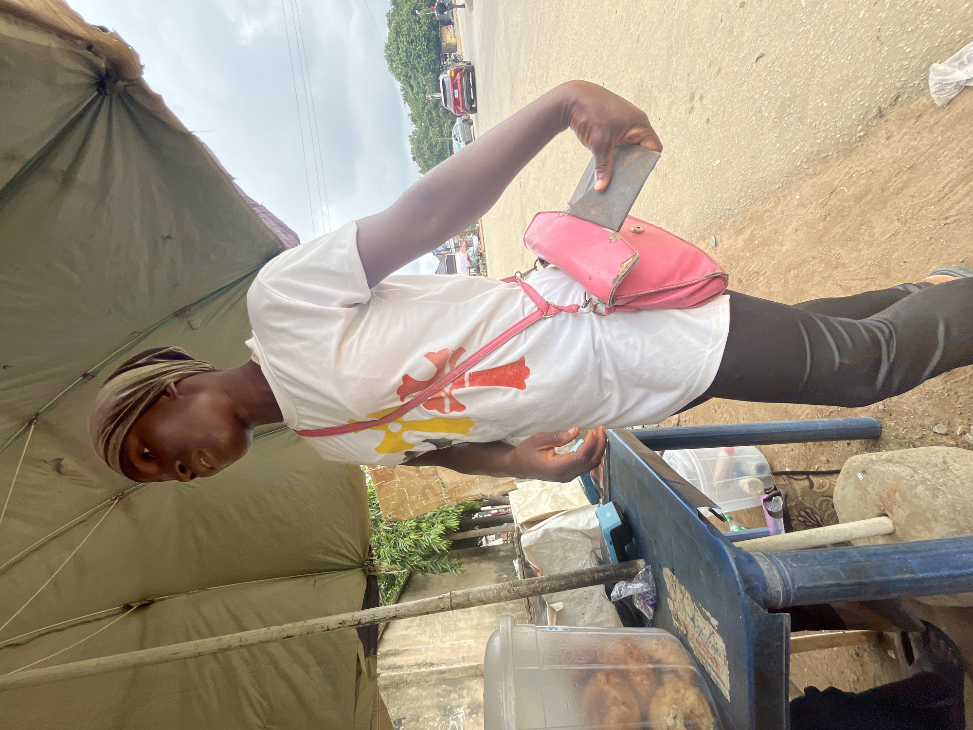 A young woman, likely a street vendor, stands under a large, weathered umbrella that provides shade. She is wearing a white t-shirt with a red, yellow, and white cross design on the front, and black leggings. A pink crossbody bag is slung over her shoulder, and a flat, rectangular object, possibly a ledger or notebook, is partially visible protruding from the flap of the bag. She is looking to her right, with a slight frown. To her left is a blue plastic table laden with items for sale. A clear plastic container filled with what appear to be fried food items sits on the table, alongside a small blue weighing scale. Several plastic bags and other assorted items are scattered on and around the table. The ground beneath the table is dirt and gravel. In the background, a paved road is visible with a few vehicles, including a red SUV, and trees line the street. The sky is overcast. The overall scene suggests a typical street market environment in Abuja, Nigeria, where vendors operate outdoors.