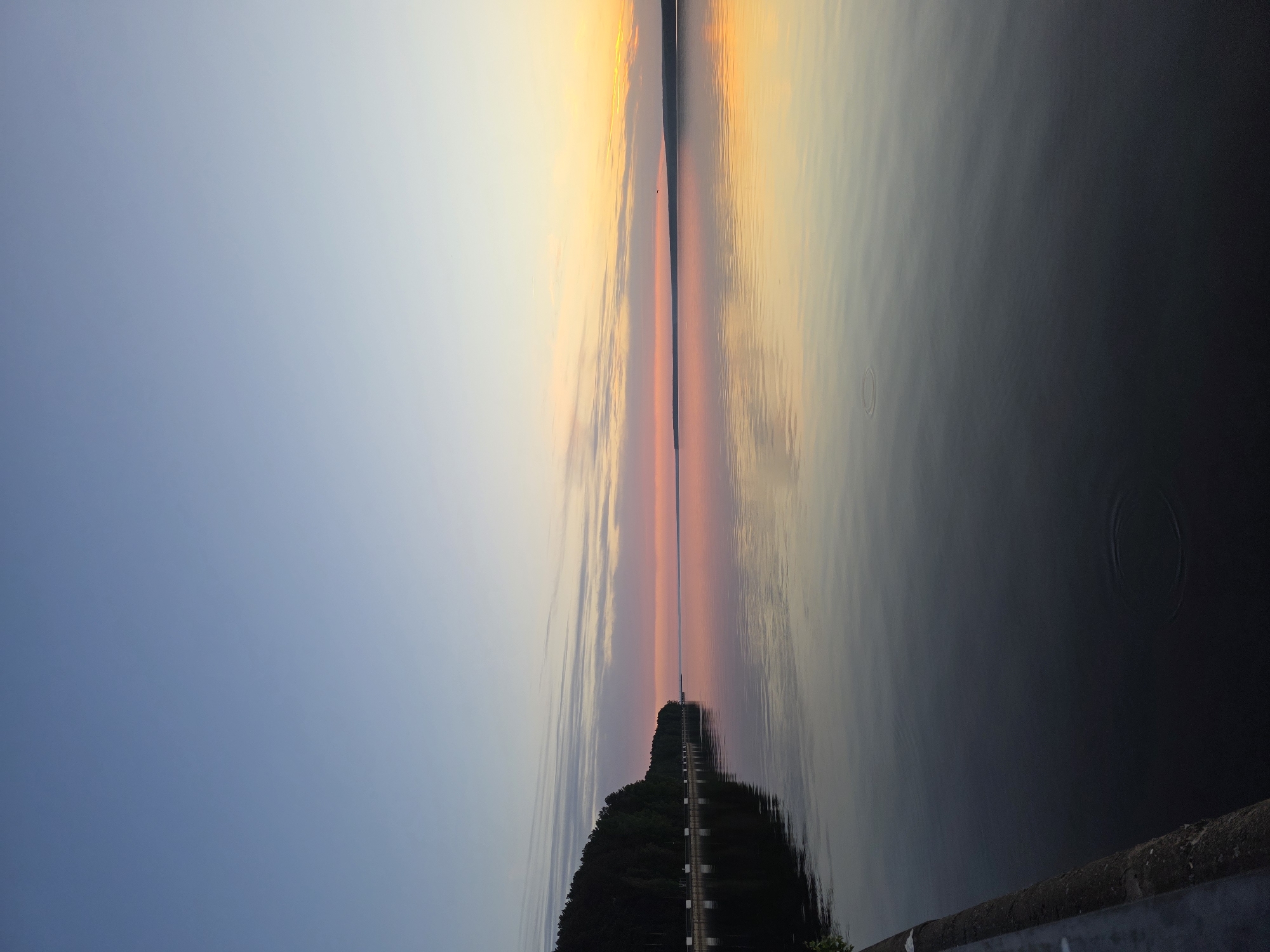 The image depicts a body of water, likely a lake or river, reflecting the sky at sunset or sunrise. A thin strip of land runs vertically through the center, suggesting a causeway or a narrow peninsula with a road. On the left side of the image, the sky transitions from a light blue to a pale yellow or orange. The right side reflects the sky, with some ripples in the water. The land area in the lower part of the image is green with a structure on it and a road running along the peninsula. The ripples in the water indicate some level of activity or disturbance. The image, is in the city of Chaikovsky, Russia.