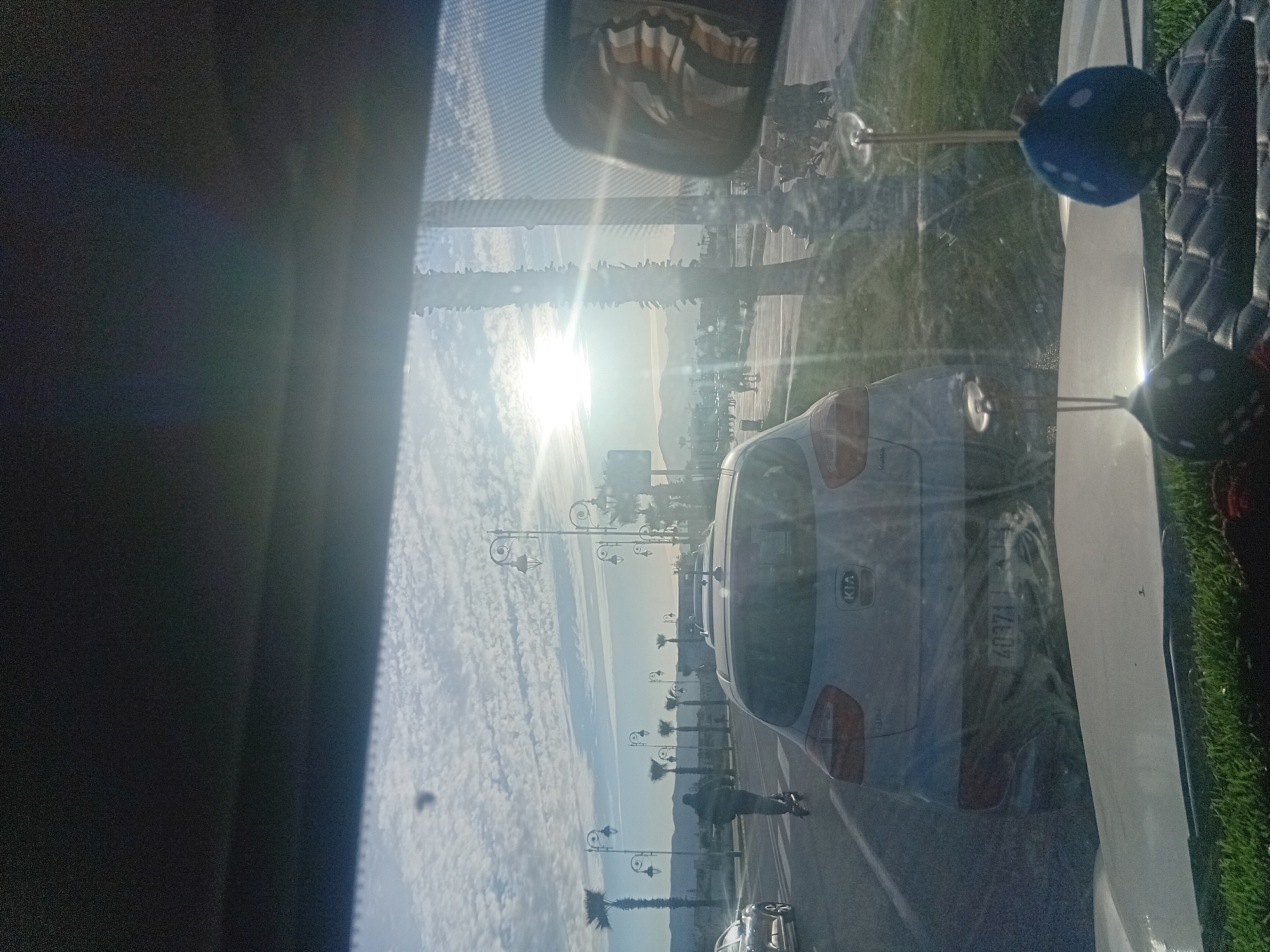A view from inside a vehicle captures a street scene in Martil, Morocco. A Kia car is visible through the windshield, parked alongside a road lined with palm trees and decorative street lamps. A person is walking on the sidewalk. The sun is shining brightly in the distance, creating a glare. The scene includes elements of urban infrastructure and a pedestrian walking along the street.