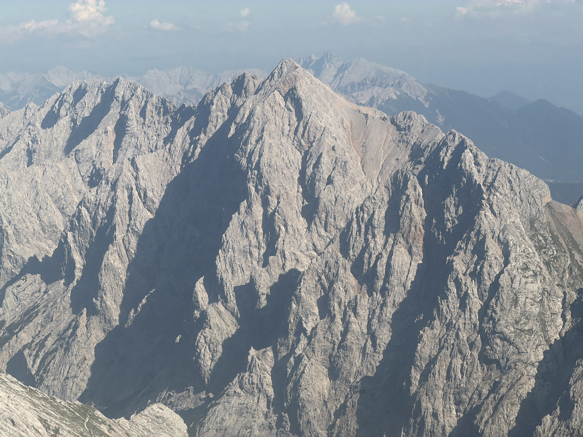 This is an aerial photograph of a rugged mountain range under a partly cloudy sky. The dominant features are the sharp, rocky peaks and deep shadows cast by the sun. The mountains are predominantly gray and brown, with some patches of green vegetation visible on the lower slopes and in shaded crevices. The overall impression is one of a wild and untamed natural landscape.
The image is a high-angle shot, suggesting it was taken from a helicopter, drone, or a much higher vantage point within the mountains themselves. The focus is on the geological formations and the play of light and shadow across the terrain. There are no signs of human habitation, infrastructure, or activity visible in the image.
This specific mountain range is likely part of the Alps. While Garmisch-Partenkirchen is a city located in the Bavarian Alps, and it is known for its mountainous surroundings including the Zugspitze, this image does not contain any specific landmarks or contextual information that definitively places it within the city's immediate vicinity or identifies a particular peak. It depicts a general mountainous landscape consistent with the region.