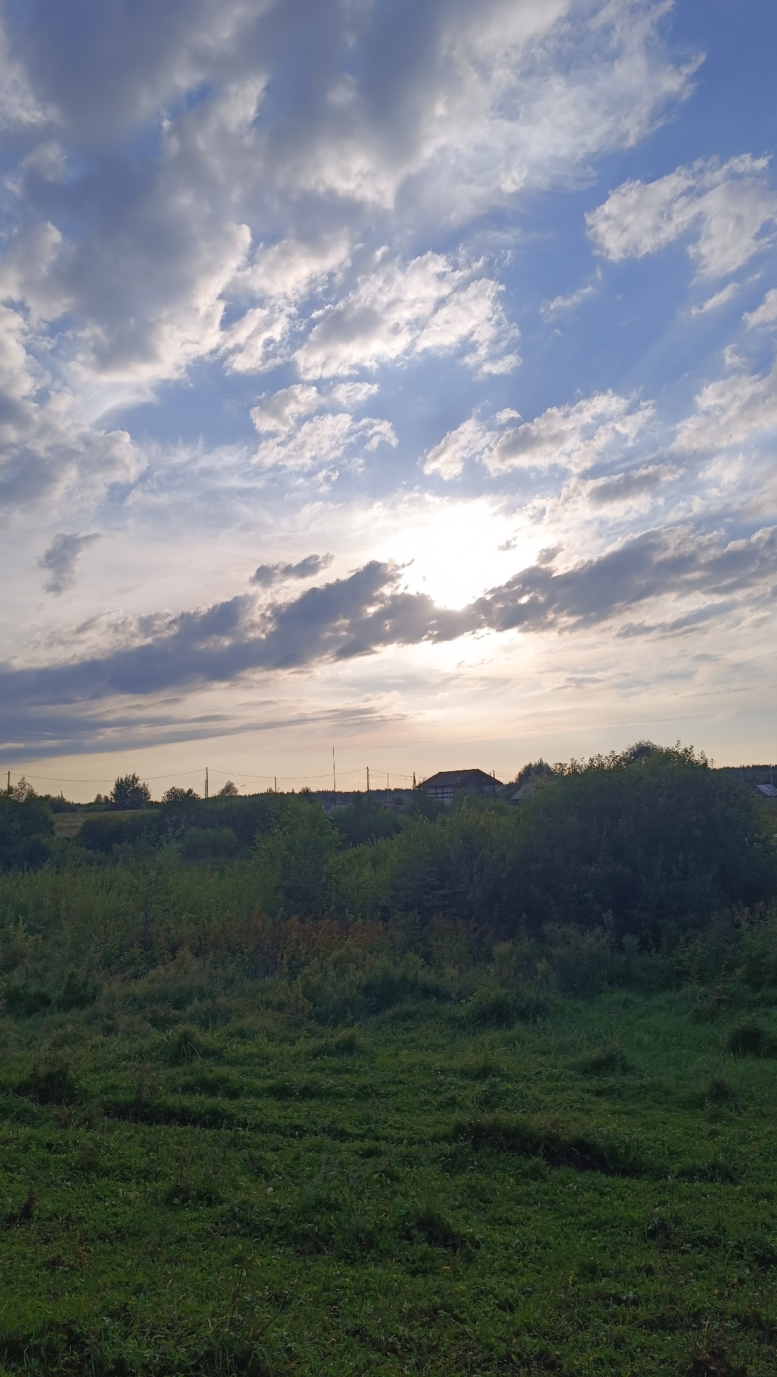 This is a vertical, eye-level, full shot image of a rural landscape under a cloudy sky. The sun is partially obscured by clouds, casting a soft, diffused light across the scene. The sky is a mix of blue and white, with elongated, layered clouds stretching horizontally across the frame. Below the sky, a dark green line of trees and bushes forms a dense barrier. In the distance, a dark-roofed house with visible windows and a section of a building can be seen peeking through the trees. Further back, a line of utility poles stretches across the horizon. The foreground is dominated by a field of green grass, some of it appearing slightly uneven or mowed. The overall impression is of a peaceful, natural environment.