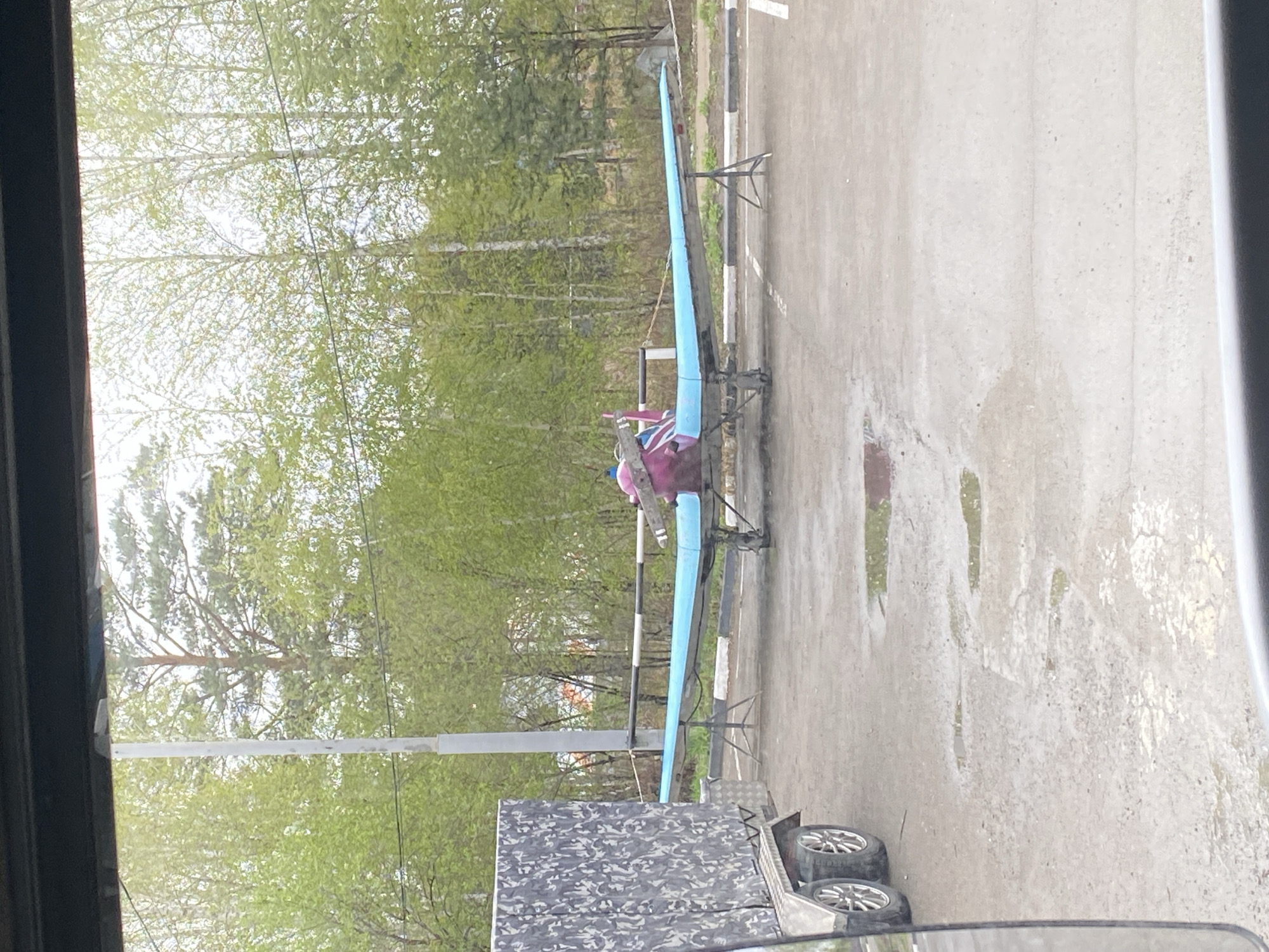 The image depicts a hang glider atop a trailer, parked on an asphalt surface. The surroundings consist of a treeline and a utility pole. The location is identified as Молодежный, Russia.