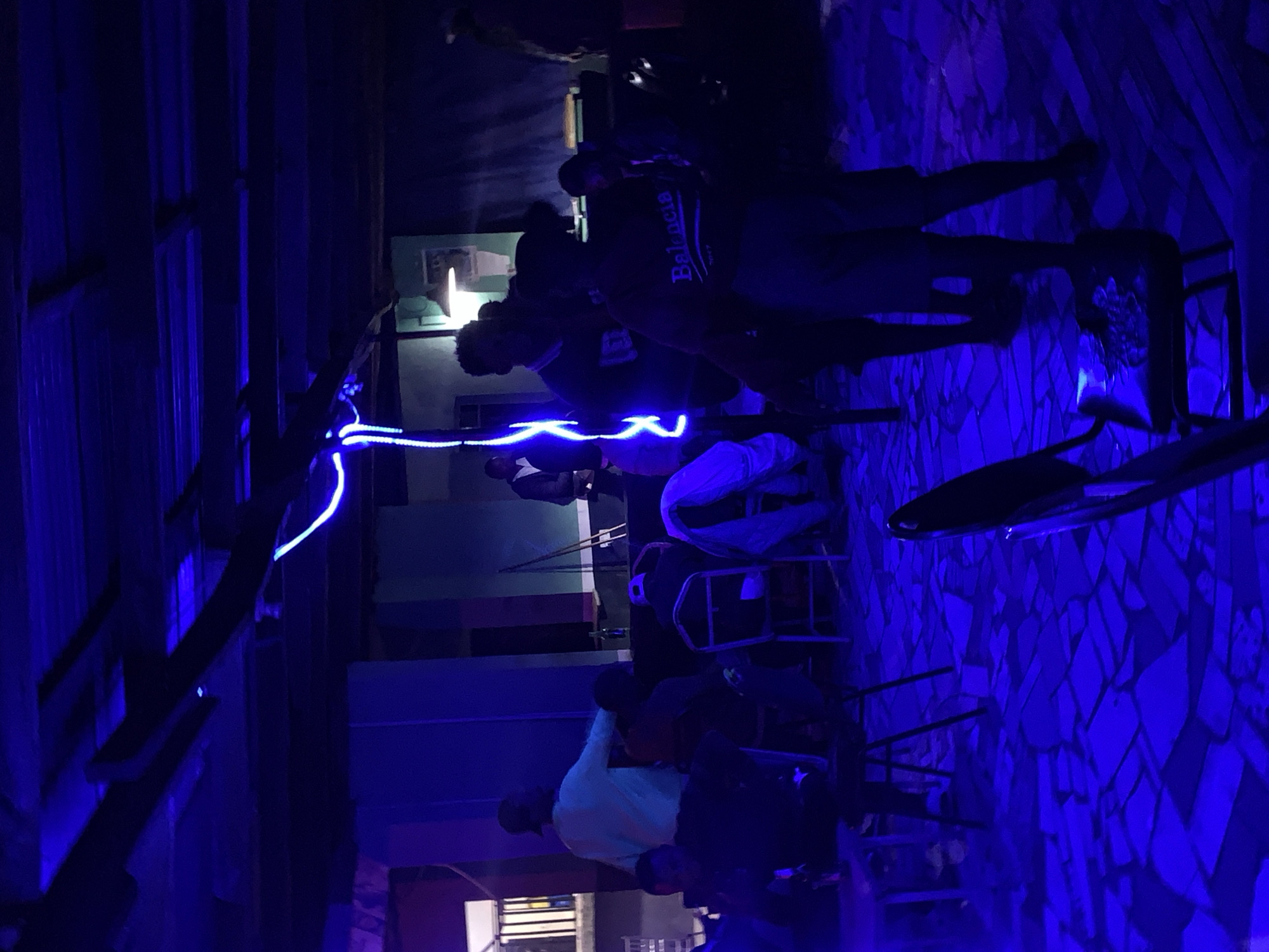 The image depicts an indoor gathering space, likely a lounge or bar, in New Stadium, Nigeria, illuminated predominantly by blue light sources, creating a low-light, monochromatic ambiance. The ceiling reveals exposed wooden beams and corrugated metal sheets. A prominent wavy blue neon or LED light strip descends diagonally from the upper right portion of the frame towards the center. The floor is covered with a mosaic pattern of irregularly shaped, flat tiles.
Multiple individuals are present, largely silhouetted or dimly lit. In the foreground-right, a person with short hair is seen from behind, wearing a dark t-shirt with "Balencia" printed on the back, and shorts. Adjacent to them, another individual stands, partially obscured, near a dark-framed chair. In the mid-ground, a person in a light-colored long-sleeved shirt and cap faces towards the left, appearing to interact with a seated individual. To the right of the center, a person wearing a patterned dark shirt is visible. Scattered throughout the scene are several dark-framed chairs, some occupied, some empty. One chair in the mid-ground-right has a white garment draped over it. In the background, a conventional light fixture with a warm-toned bulb is visible on a light green wall. Further back, a dark counter or bar-like structure is present, with individuals standing near it. A bottle is visible on a surface in the mid-ground. The general activity appears to be social gathering, with individuals standing and sitting, engaging in conversation or waiting.