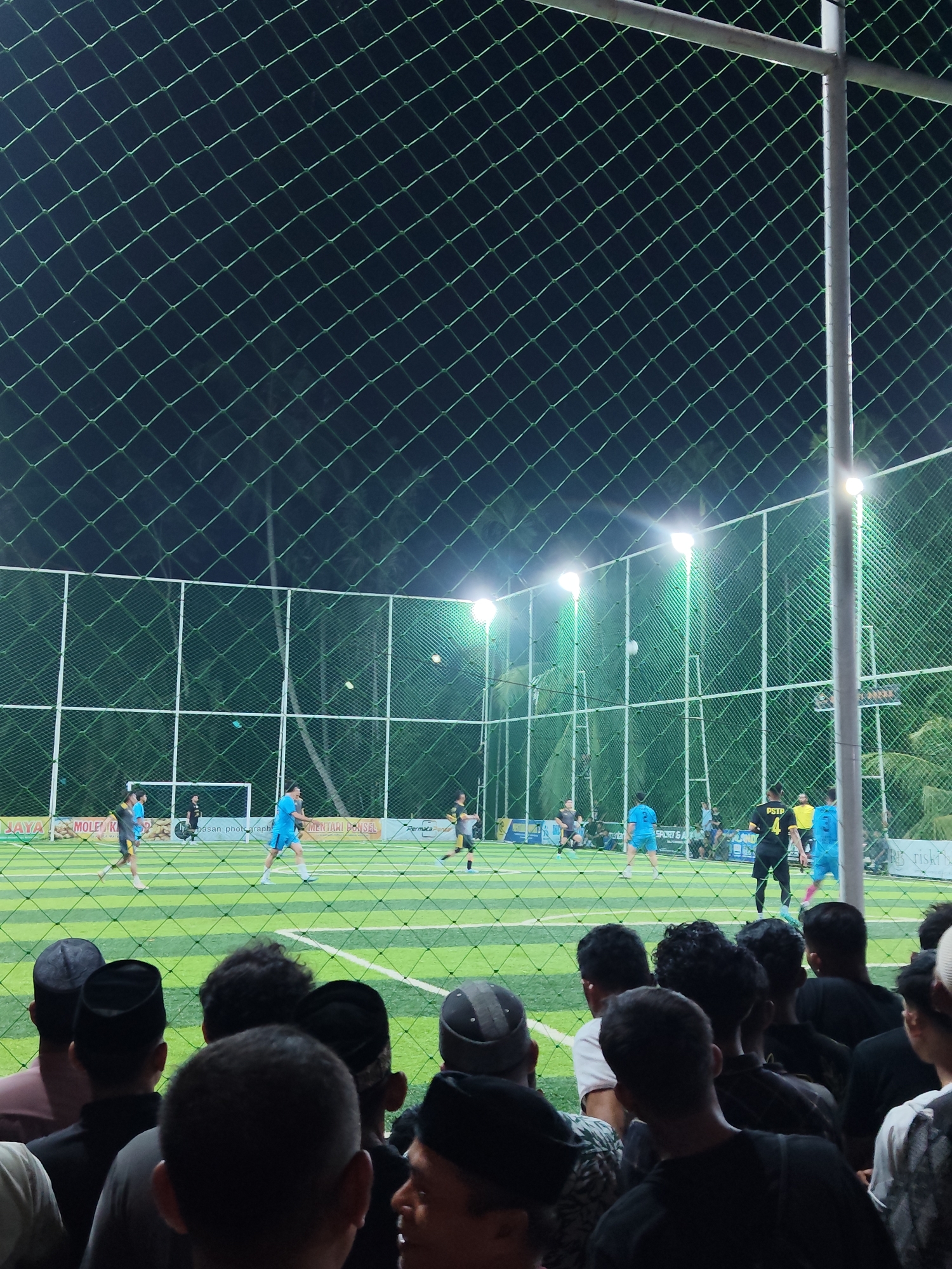 The image depicts an outdoor futsal court at night, illuminated by floodlights, located in Mee, Indonesia. The court's artificial turf is striped green and white. A net enclosure surrounds the court, with a green mesh visible in the foreground, partially obscuring the view.
On the court, several men are playing futsal. They are wearing athletic attire, including jerseys and shorts in various colors such as blue, grey, and yellow. Some players are actively engaged in the game, with one player in a blue jersey running with the ball, and others positioned on the field. A goal is visible at the far end of the court.
Beyond the court, a crowd of spectators is gathered behind the net. These individuals are mostly men, and many are wearing traditional skullcaps (peci). They are observing the game, with some looking towards the players and others interacting with each other. The background shows palm trees and distant structures, characteristic of an Indonesian landscape. The overall atmosphere appears to be one of community engagement and sporting event.