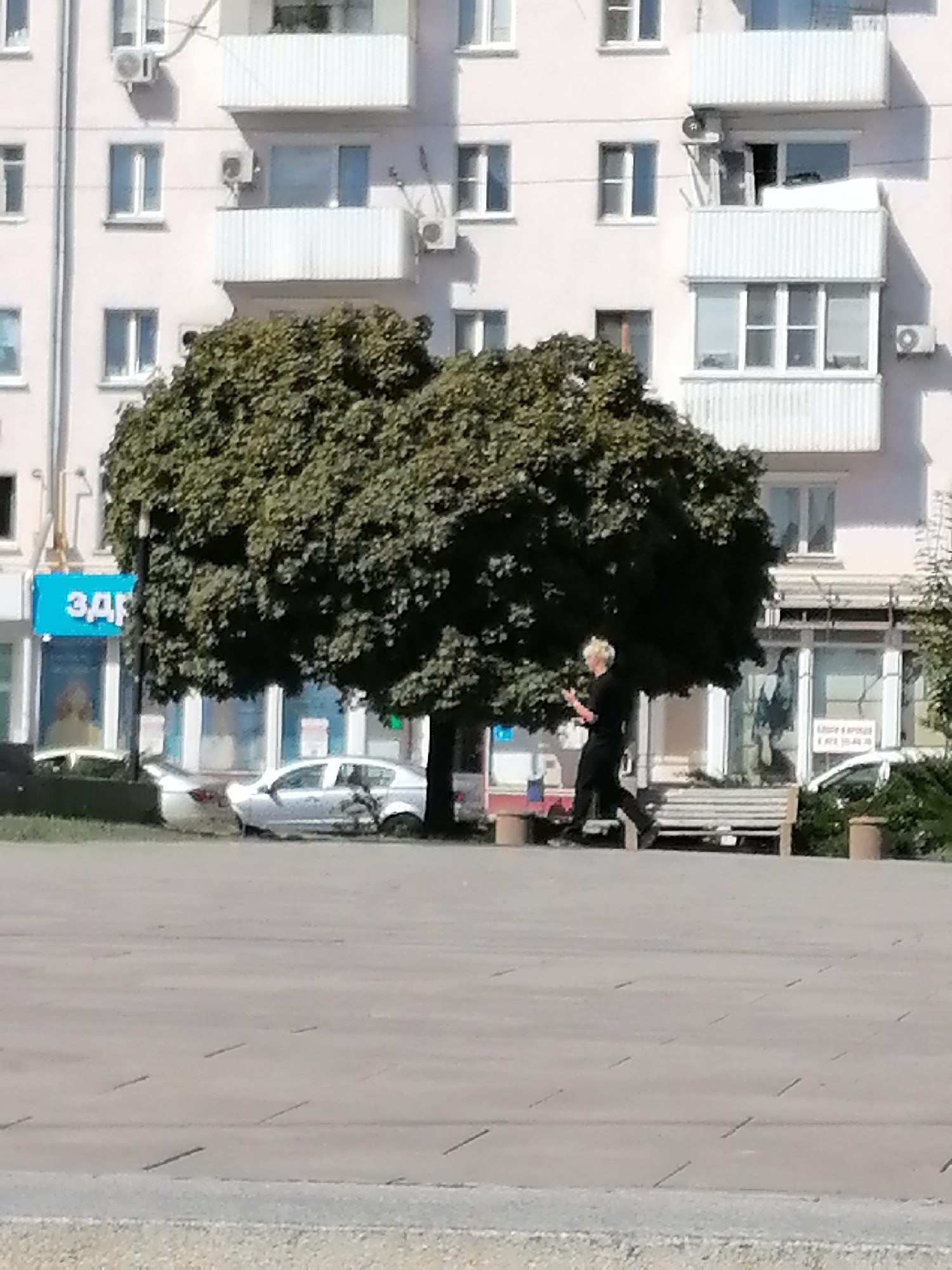 In Rostov-on-Don, Russia, a person with blonde hair and dark clothing is walking past a large green tree in front of a building. The building appears to be an apartment complex with multiple floors and balconies. A silver car is parked on the street in front of the building, partially obscured by the tree. There is a park bench to the right of the tree. The scene is set in an urban environment with pavement in the foreground and a building and tree in the background.