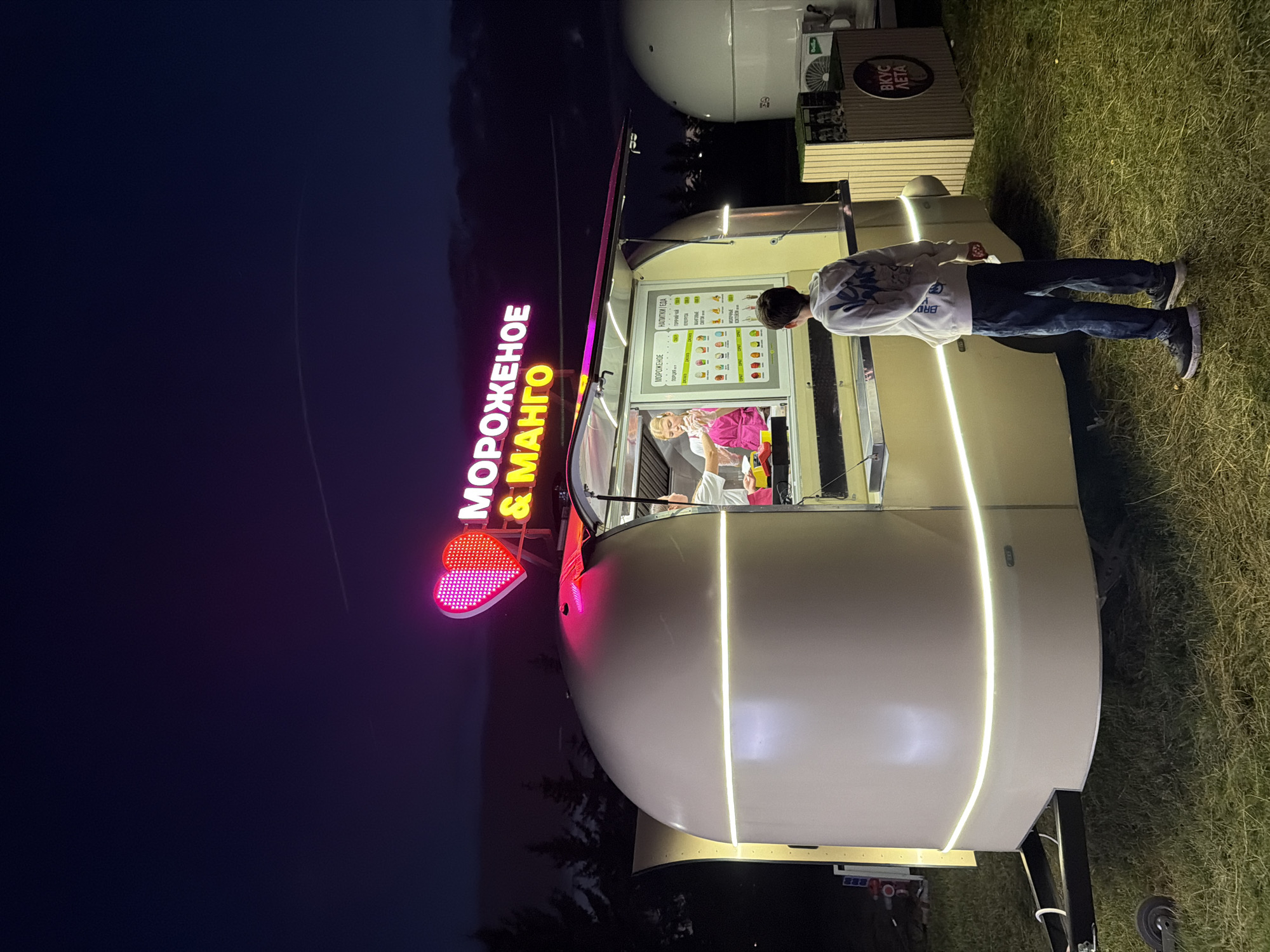 At night, a young boy in a white hoodie and dark pants stands facing a food truck. The food truck is a white, rounded trailer with illuminated strips of light. Above the service window, a neon sign in pink and yellow displays the words "МОРОЖЕНОЕ & МАНГО" (Ice Cream & Mango) with a pink heart icon. Inside the open service window, a person in a pink shirt is visible. A menu board inside the truck shows various ice cream and drink options. To the right of the truck, a wooden stand with a sign reading "ВКУС ЛЕТА" (Taste of Summer) is visible. The ground is covered in green grass, and trees are silhouetted against the dark sky in the background. A faint contrail is visible in the night sky.