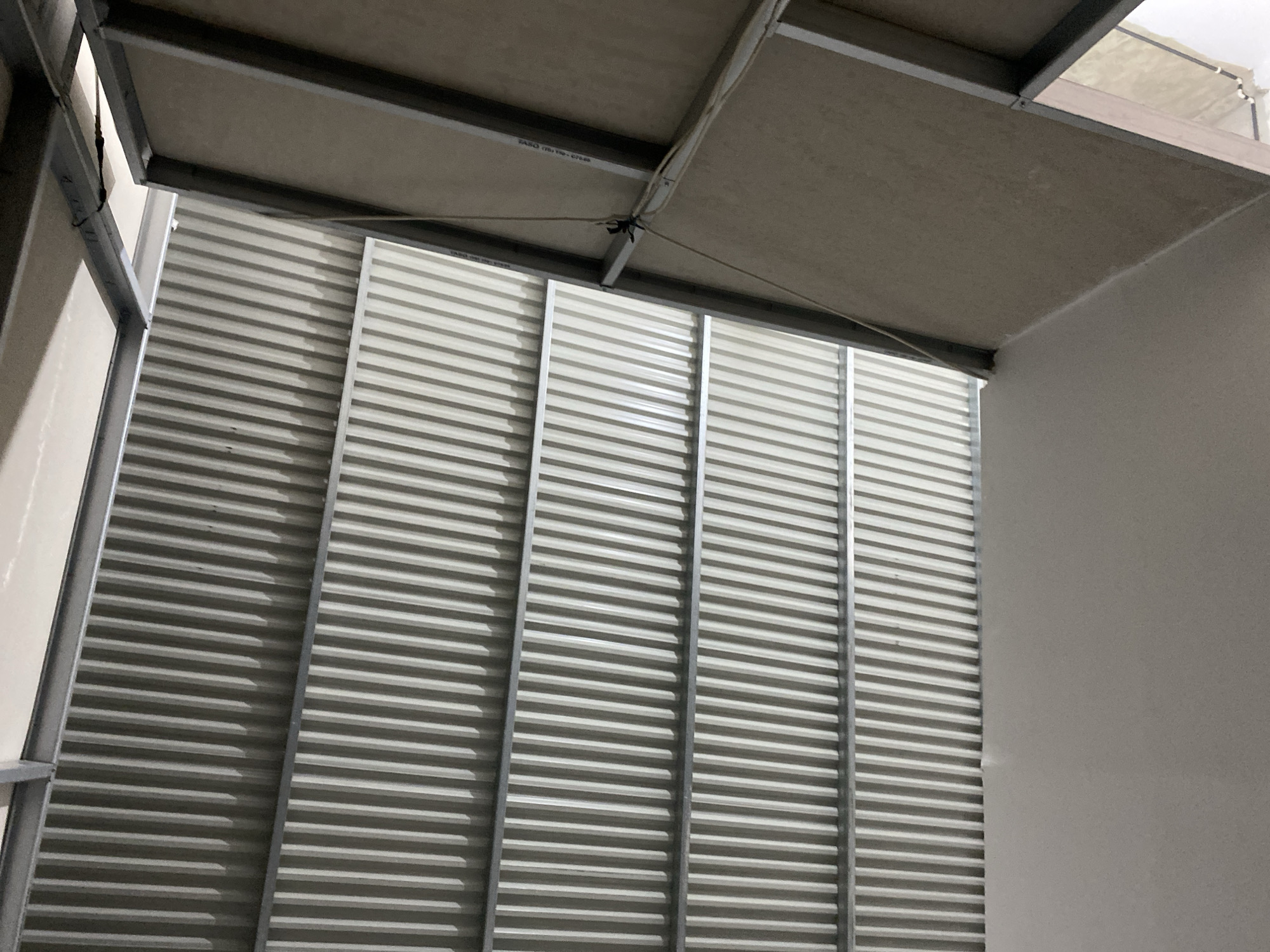 A low-angle, full shot captures the underside of a corrugated metal roof structure, seemingly under construction or renovation. The ribbed, metallic ceiling panels are supported by horizontal metal beams. To the right, a section of a tan, textured wall is visible, also appearing unfinished or exposed. A thin, light-colored rope or wire dangles from the upper beams towards the wall. The lighting is dim, suggesting an indoor or covered space with minimal direct light. The scene is devoid of people or any specific identifiable markers of location within Ta', Indonesia.