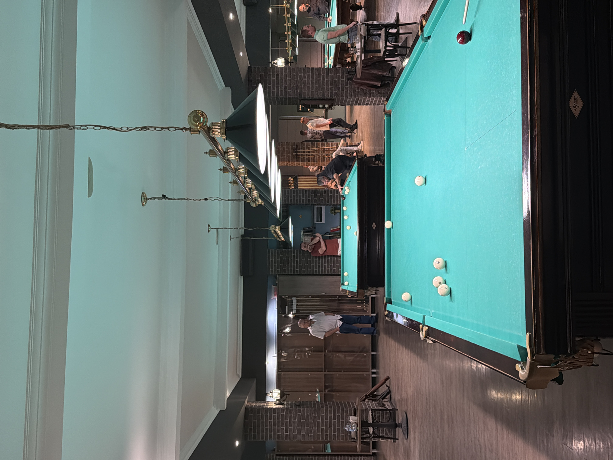 This image depicts the interior of a billiard hall located in Izhevsk, Russia. The establishment features multiple billiard tables with vibrant green felt surfaces, well-illuminated by rows of ornate brass-colored pendant lamps with dark, conical shades suspended from the ceiling by chains. The ceiling itself is a combination of white panels with decorative molding and dark gray sections, exhibiting a faint greenish-blue tint in areas, possibly due to lighting or reflection.
The walls are characterized by a mix of exposed reddish-brown brickwork on structural columns and lower sections, transitioning to dark gray painted surfaces above. The flooring consists of light-colored wooden planks. Along one wall, a series of dark wooden locker units are visible, suggesting storage for personal belongings or billiard equipment. Wall sconces with frosted glass shades provide additional ambient lighting.
Several adult males are present throughout the hall. In the foreground, a billiard table displays four white balls and one dark red object ball, with a billiard cue positioned near the red ball. Another table further back is also in play. Men are engaged in various activities: some are actively playing billiards, leaning over tables with cues, while others are standing and observing. Specifically, a man in a white collared shirt and blue jeans stands with his hands in his pockets near the locker units on the left. Another man in a red T-shirt leans against a brick column on the right side of the image. A man in a light green polo shirt stands near a billiard table on the right, holding a cue. Small, dark metal tables with chairs are placed around the hall for seating, one with a water bottle on it. The logo "Старт" (Start) is visible on the side panel of the closest billiard table.