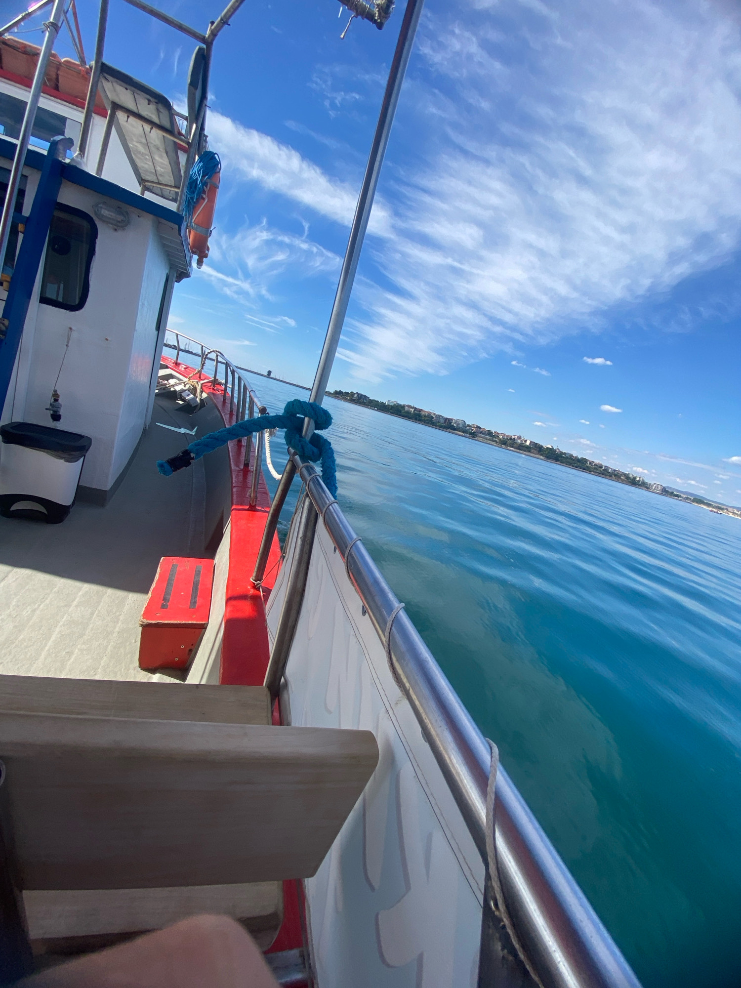 The image shows a boat on the water near the city of Nesebar, Bulgaria. The boat is white with a red railing and blue trim. There is a life preserver and a blue rope on the deck of the boat. The water is blue and calm, and the sky is clear with some white clouds. The city of Nesebar is visible in the background, with buildings and trees lining the coast. The perspective is from the deck of the boat, looking out towards the water and the city. There is a wooden bench visible in the foreground.