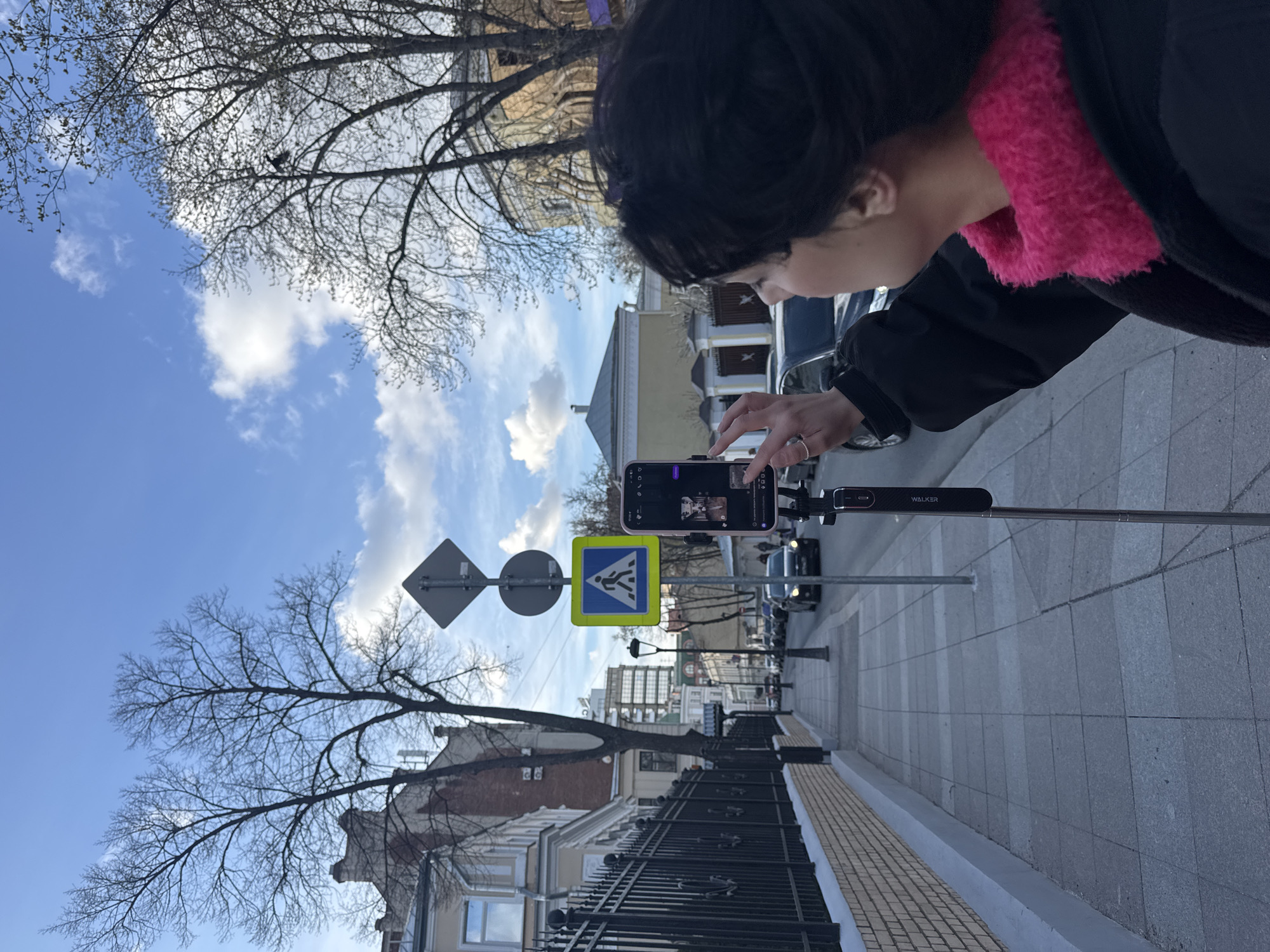 A person with dark hair and a pink collar is operating a smartphone mounted on a "WALKER" branded selfie stick on a sidewalk. The sidewalk consists of grey stone tiles, and a fence with a light stone base runs along the side. Beyond the fence are buildings with architectural details. The person is positioned near street signs indicating a pedestrian crossing. Further into the background is a street with parked and moving vehicles. The sky above is blue with scattered clouds. The image appears to have been taken on a street in Moscow, Russia, judging by the style of the buildings and street signage.