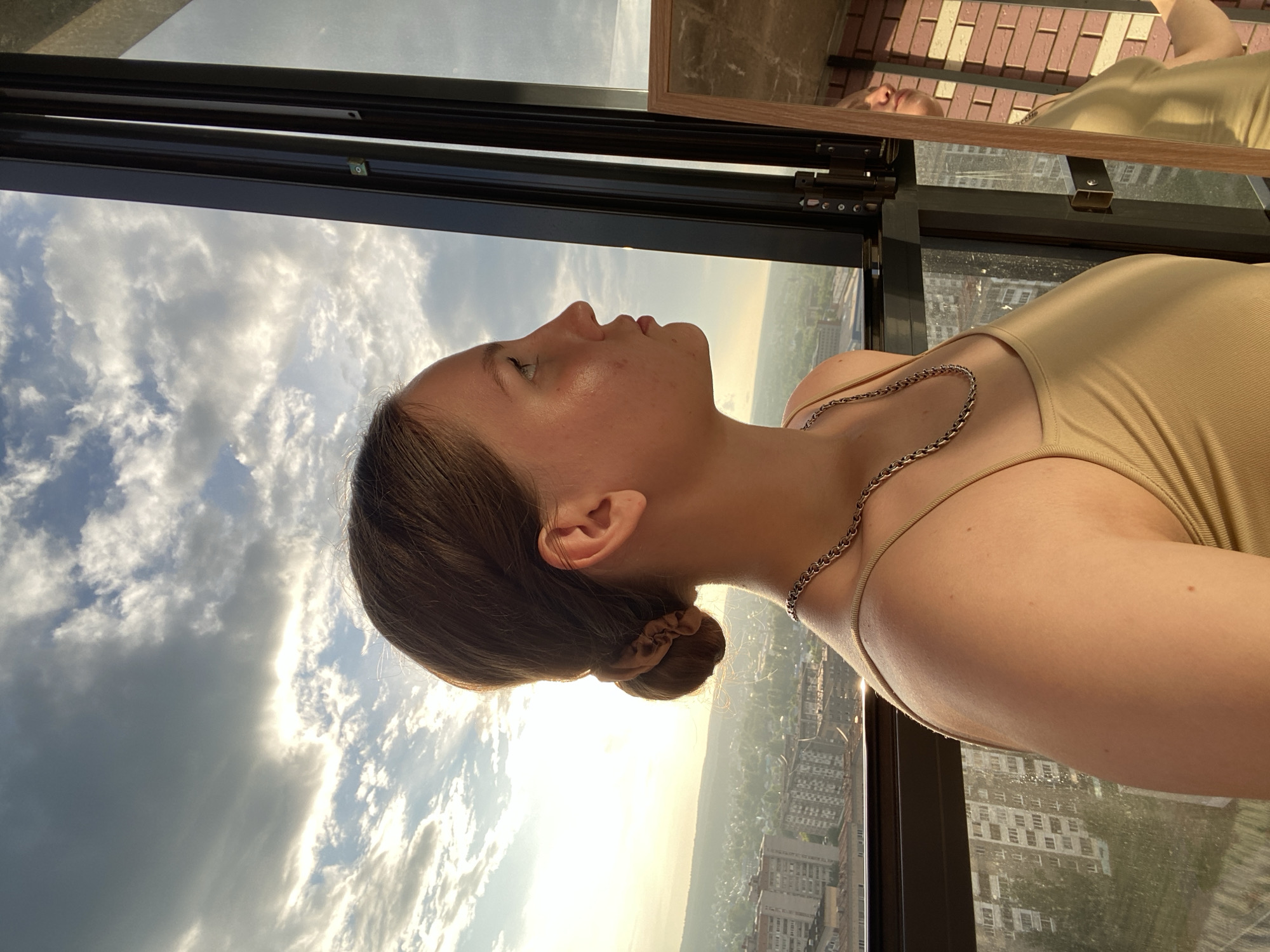 A young woman with brown hair pulled back in a bun is shown in profile, looking out a window. She is wearing a gold-colored camisole and a silver chain necklace. The background is a city skyline with buildings and a cloudy sky. The setting appears to be an apartment or office building in Izhevsk, Russia, with a mirror to the right of the woman reflecting a portion of her body and the room behind her. The lighting suggests it is either sunrise or sunset.