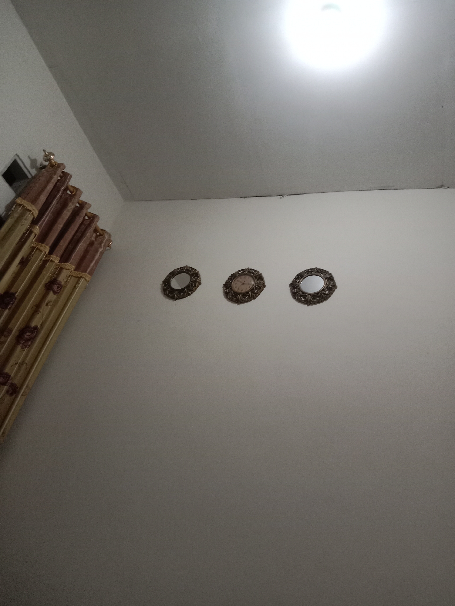 The image shows an interior view of a room, likely a living room or hallway, in Brangsong, Indonesia. The room has white walls and a white ceiling. On the wall, there are three decorative circular items. The item on the left is a clock with Roman numerals. The middle item is a mirror. The item on the right is another mirror. To the left of these items, a set of curtains hangs from a curtain rod. The curtains are ornate, with a gold and brown floral pattern. A light fixture is visible on the ceiling, casting a bright glow. The overall impression is of a modestly decorated residential interior.