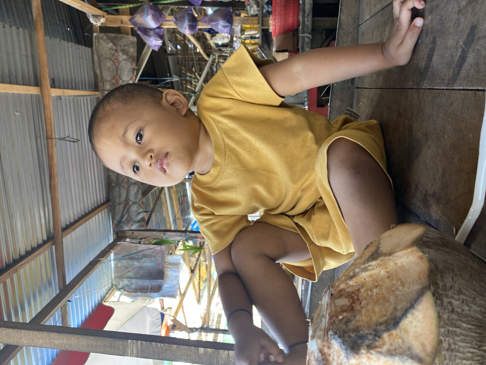 The image captures a young child, likely a toddler, sitting on a wooden surface, possibly a table or a bench, within an open-air structure. The child is wearing a mustard-yellow t-shirt and shorts. Their left leg is bent, with the knee pointing upwards, while the right leg is extended forward. The child's gaze is directed upwards and to the left. In the foreground, a partially visible coconut with a straw is present. The background reveals a rustic, possibly market-like environment with a corrugated metal roof supported by wooden beams. Various items are visible in the background, including what appear to be plastic bags hanging, shelves, and containers. The lighting suggests it is daytime, with natural light filtering into the structure. The overall impression is of a candid moment captured in a humble, everyday setting. Based on the architecture and the presence of the coconut, the location is likely Ta', Indonesia, a region known for its tropical climate and abundant coconut production.