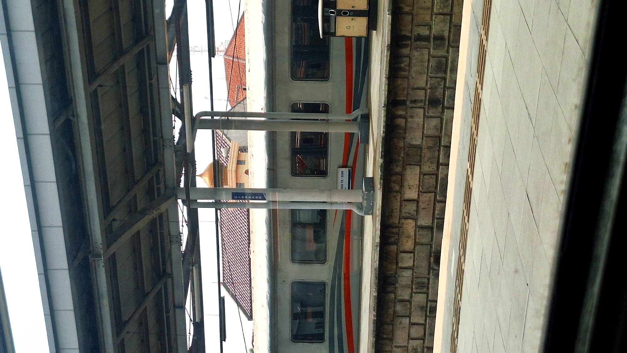 This image depicts a train at a station platform. The train is painted in a gray and white color scheme with a red stripe. It is located next to a brick wall and a tiled platform. Overhead structures support the roof of the station. In the background, a building with a tiled roof and a golden dome is visible. A sign on the train car reads "Cikarang," indicating the location in the city of Cikarang, Indonesia.