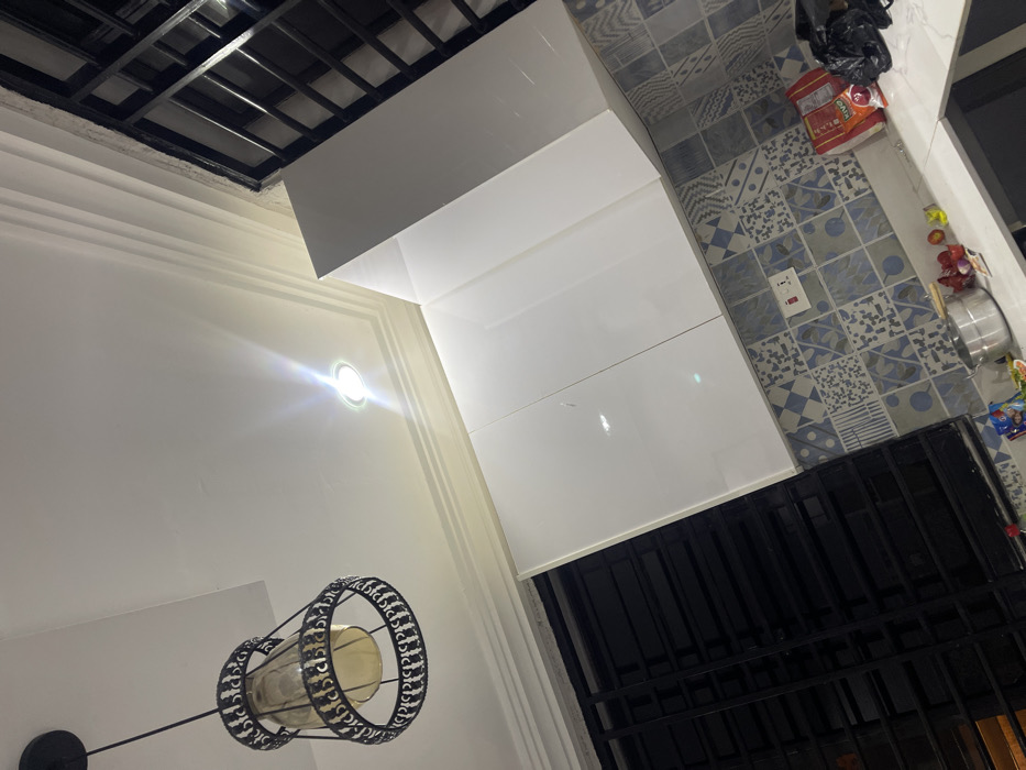 This media file captures an indoor scene, likely a kitchen or utility area, within a residence in Adenta Municipality, Ghana. The image is taken at an angle, resulting in a tilted perspective.
The room features clean white walls adorned with decorative crown molding and a white ceiling. A section of a black grid structure, possibly a decorative ceiling element or part of a suspended ceiling, is visible in the upper left. The space is illuminated by two light sources: a circular recessed light fixture emitting a bright glow on the wall, and a decorative hanging pendant light in the lower left, featuring a black metal cage surrounding a cylindrical glass shade.
A significant portion of the image is occupied by modern, glossy white L-shaped cabinetry, likely kitchen cupboards, which reflect the light. The floor or a backsplash area is covered with distinct patterned tiles in a mix of light blue, grey, and white, showcasing various geometric and abstract designs. A white electrical outlet with a switch is mounted on this tiled surface.
On the tiled surface, towards the upper right, a black plastic bag, a red and white packaged item (partially showing "KIVO" text, suggesting a brand name), and other small items are visible. Further down, a stainless steel pot or bowl and more small packaged goods are present. In the lower right, a dark, black metal-barred structure, possibly a window with security bars or a door, is seen. The prominent artificial lighting suggests the time of day could be evening or night, or that the interior requires constant illumination. No individuals are present, and no specific activity is taking place.
