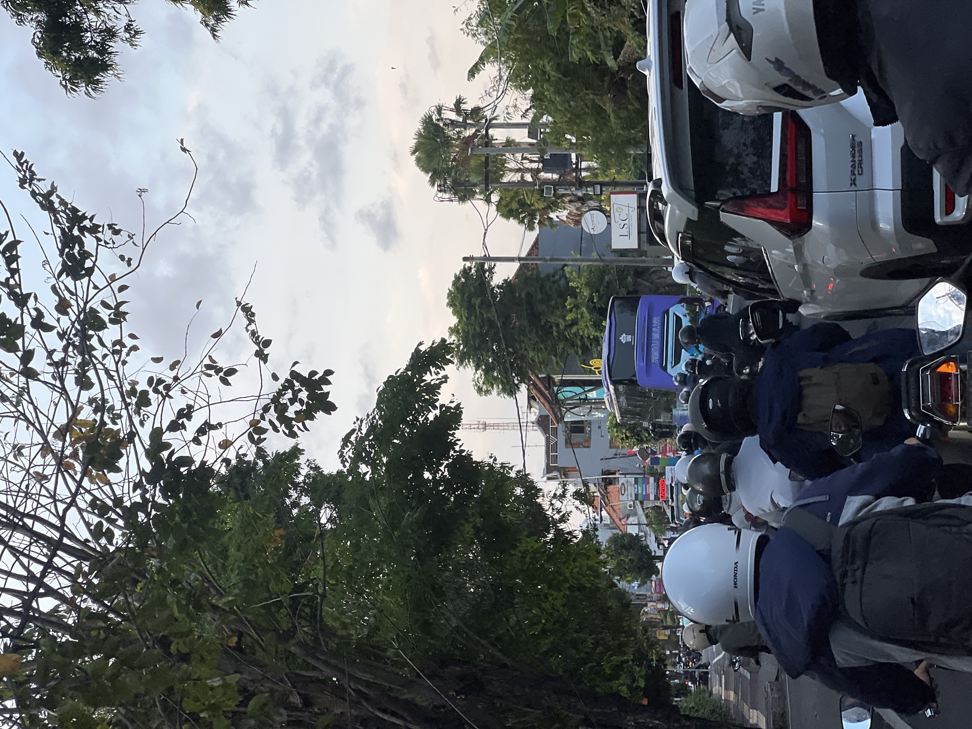 The image depicts a traffic scene in Denpasar, Indonesia. The foreground shows the backs of several motorcyclists wearing helmets. One motorcyclist in the center wears a white helmet with "HONDA" written on it and a blue jacket over a backpack. To their right, another motorcyclist wears a black helmet and a dark jacket with a backpack. To the left, a motorcyclist wears a white helmet and a grey jacket. They are all on motorcycles, positioned on a road with other vehicles.
In the mid-ground, there is a white SUV, possibly a Mitsubishi Xpander Cross, with its rear facing the viewer. To the left of the SUV is a blue bus with "FESTU MULLA" written on its side. Behind the bus, there are buildings with signs, including one with "LSC" prominently displayed. Power lines and utility poles are visible, with palm trees and other foliage interspersed with the urban structures.
The background shows a continuation of the traffic, with more motorcycles and cars, and buildings receding into the distance. The sky is overcast with soft clouds, suggesting either early morning or late afternoon. The overall impression is of a busy street in Denpasar.