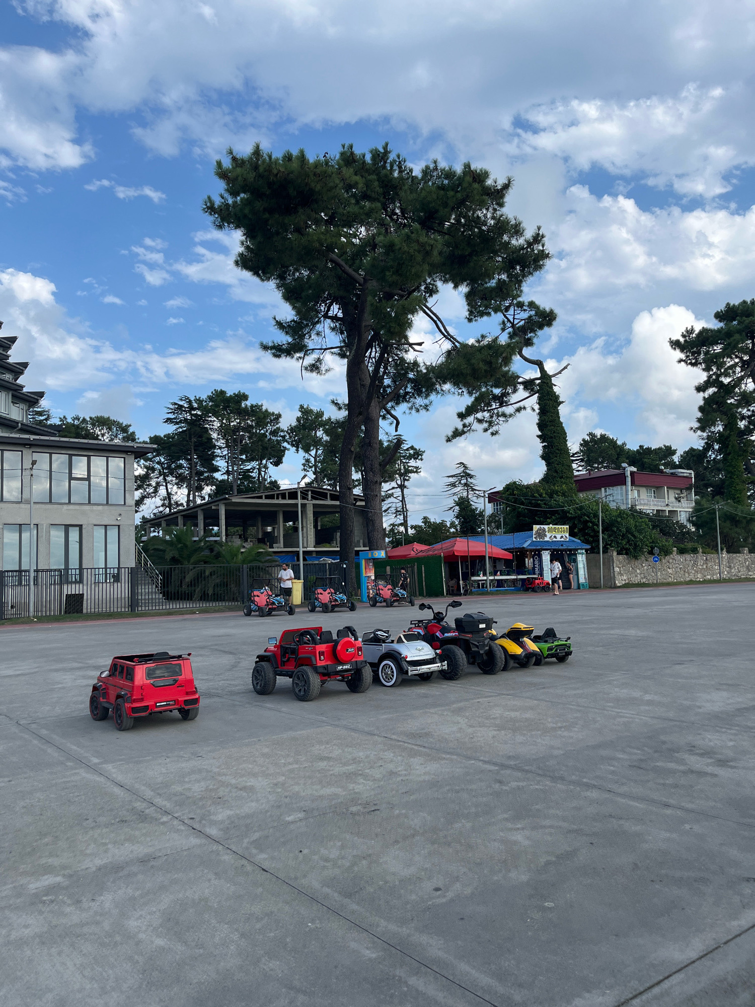 In the city of Ureki, Georgia, a paved outdoor area is filled with children's electric ride-on vehicles. In the foreground, two red toy jeeps are parked side-by-side. Behind them, a line of more toy vehicles includes a silver classic car, a red ATV, a yellow ATV, and a green ATV. In the background, a large, old pine tree dominates the scene, with its branches extending across the sky. To the left, a modern, multi-story building with large windows stands beside a more open, partially constructed building. Several small kiosks with red and blue awnings are visible further back, with a few people standing near them. The sky above is a bright blue with scattered white clouds.