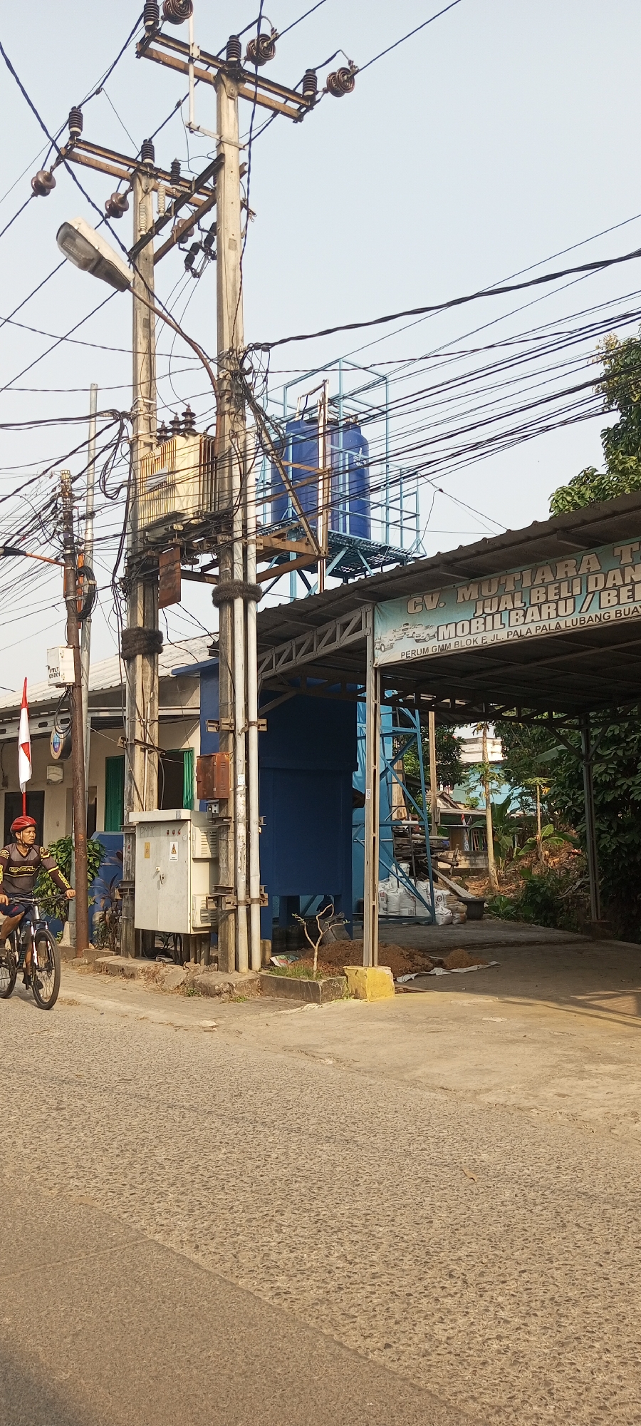 The image captures a street scene in Setu, Indonesia, on a clear, bright day. In the foreground, a man wearing a red helmet is riding a bicycle on a paved road with a textured surface. He is dressed in a dark long-sleeved shirt with yellow accents and dark shorts. To the left of the road, a white electrical transformer box is mounted on a utility pole that is heavily adorned with a dense network of electrical wires. Further back, a large blue water tank is elevated on a metal structure. Adjacent to this, a building with a dark roofed overhang displays a banner with Indonesian text that appears to advertise a business related to car sales. Behind the building, lush green trees and vegetation are visible. The environment suggests a mix of urban infrastructure and natural elements.
