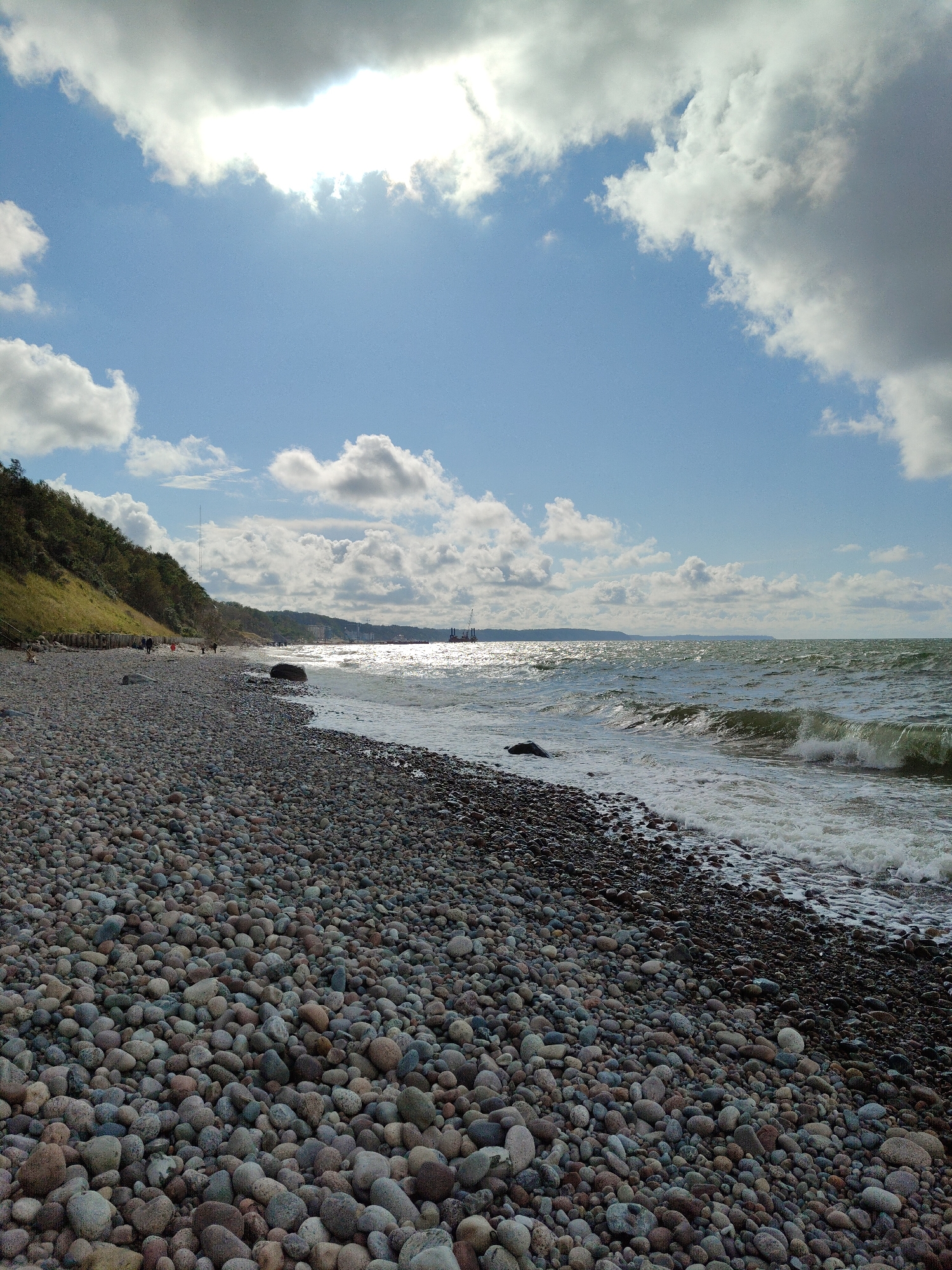 This is a vertical, eye-level, full shot of a pebble beach on the Baltic Sea coast in Pionersky, Russia, under a partially cloudy sky. The foreground is dominated by a dense scattering of smooth, rounded pebbles in various shades of gray, brown, and pink. A gentle wave breaks at the edge of the water, creating white foam that washes onto the shore.
In the midground, the sea stretches out to the horizon, reflecting the sunlight which creates a shimmering effect. Beyond the sea, a faint coastline is visible, with what appear to be industrial structures or cranes on the far right.
To the left, a steep, grassy hill slopes down towards the beach, dotted with trees and shrubs. A wooden walkway or stairs can be seen on the hillside. Several individuals are present on the beach and along the edge of the hill, appearing as small figures walking or standing. The sky above is a mix of bright blue and large, fluffy white and gray clouds, with the sun partially obscured by a large, bright cloud formation in the upper left quadrant.