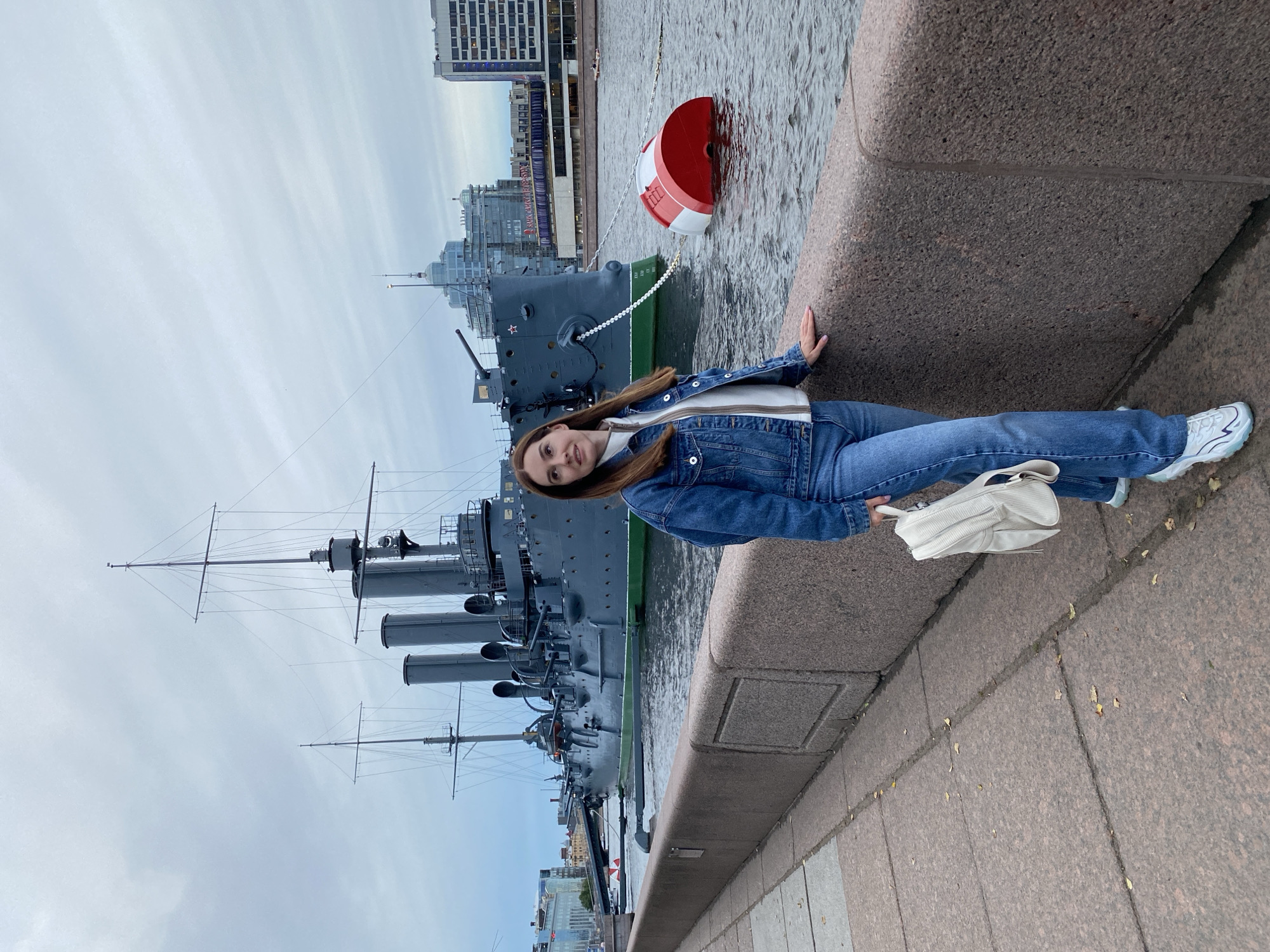 A young woman with long brown hair is sitting on a stone embankment, facing the camera. She is wearing a denim jacket, a white shirt, and blue jeans. Her right hand rests on the stone beside her, and she holds a white purse in her left hand. She is smiling, and her expression suggests a relaxed and happy mood.
Behind her, a large, dark grey ship is docked in a body of water. The ship has multiple smokestacks and tall masts, suggesting it is a historical vessel. A red and white buoy is floating in the water to the right of the ship. In the background, modern buildings with glass facades are visible along the waterfront. The sky is overcast and cloudy, indicating a cool, possibly autumnal day.
The scene appears to be a tourist spot in Saint Petersburg, Russia, likely a waterfront area where historical ships are displayed or docked. The woman's casual attire and the presence of a tourist attraction suggest she is a visitor enjoying the sights. The overall atmosphere is calm and serene. There is no visible text in the image.