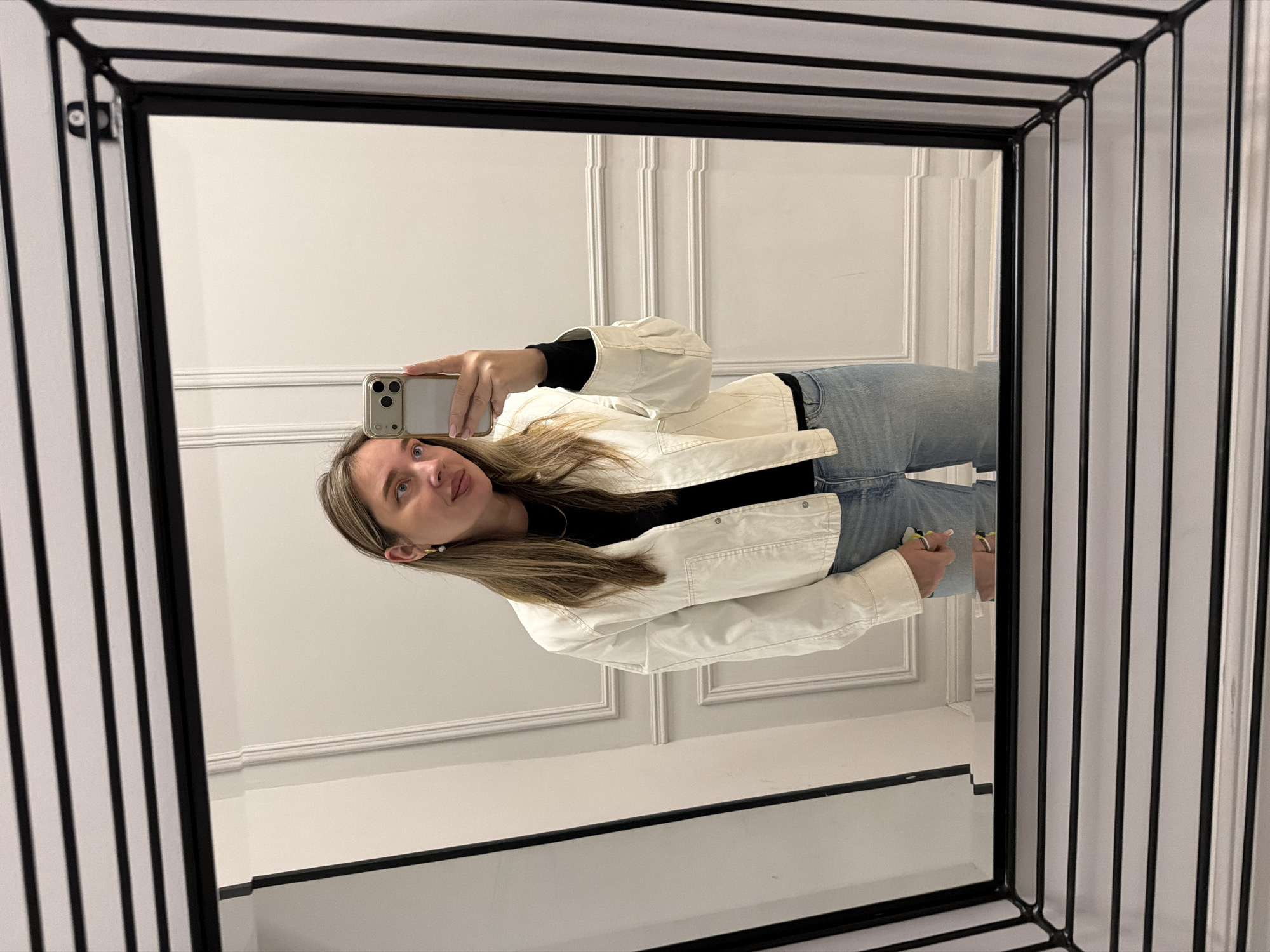 A young woman captures her reflection in a mirror, taking a mirror selfie with her phone. She is positioned slightly to the left, holding a light-colored smartphone with a visible camera array in her right hand, aimed at the mirror. She has long, light brown hair, a neutral expression with slightly parted lips, and a small earring in her visible ear. She is dressed in a black top, a light-colored (possibly cream or off-white) jacket with noticeable stitching and buttons, and light blue denim jeans.
The mirror she is reflecting in features a unique, thick, black geometric frame composed of multiple parallel lines and angles, creating a sense of depth around the reflective surface. The background visible within the mirror's reflection is a plain, light-colored wall (likely white or off-white) adorned with decorative wainscoting or paneling, characterized by raised moldings. The overall setting is indoors, with soft, even lighting, suggesting a well-lit interior space in Moscow, Russia. No visible text is legible in the image.