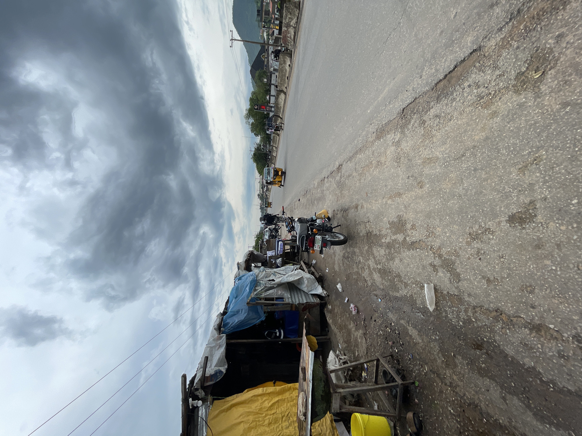 The image depicts a roadside scene in Lokoja, Nigeria, with a focus on a makeshift stall selling various items. The stall is constructed from corrugated metal and covered with tarps of blue, white, and yellow, suggesting it's a temporary or informal structure. Several motorcycles are parked in front of the stall, and a yellow tricycle is visible on the road. In the background, a paved road stretches into the distance, with some buildings and a hill on the left side. Power lines are visible overhead. The sky is overcast with dark, heavy clouds, indicating a cloudy or potentially rainy day. The lighting suggests it could be late afternoon. The scene appears to be a busy commercial area, with evidence of pedestrian activity and parked vehicles. There is some litter scattered on the ground around the stall and on the roadside.