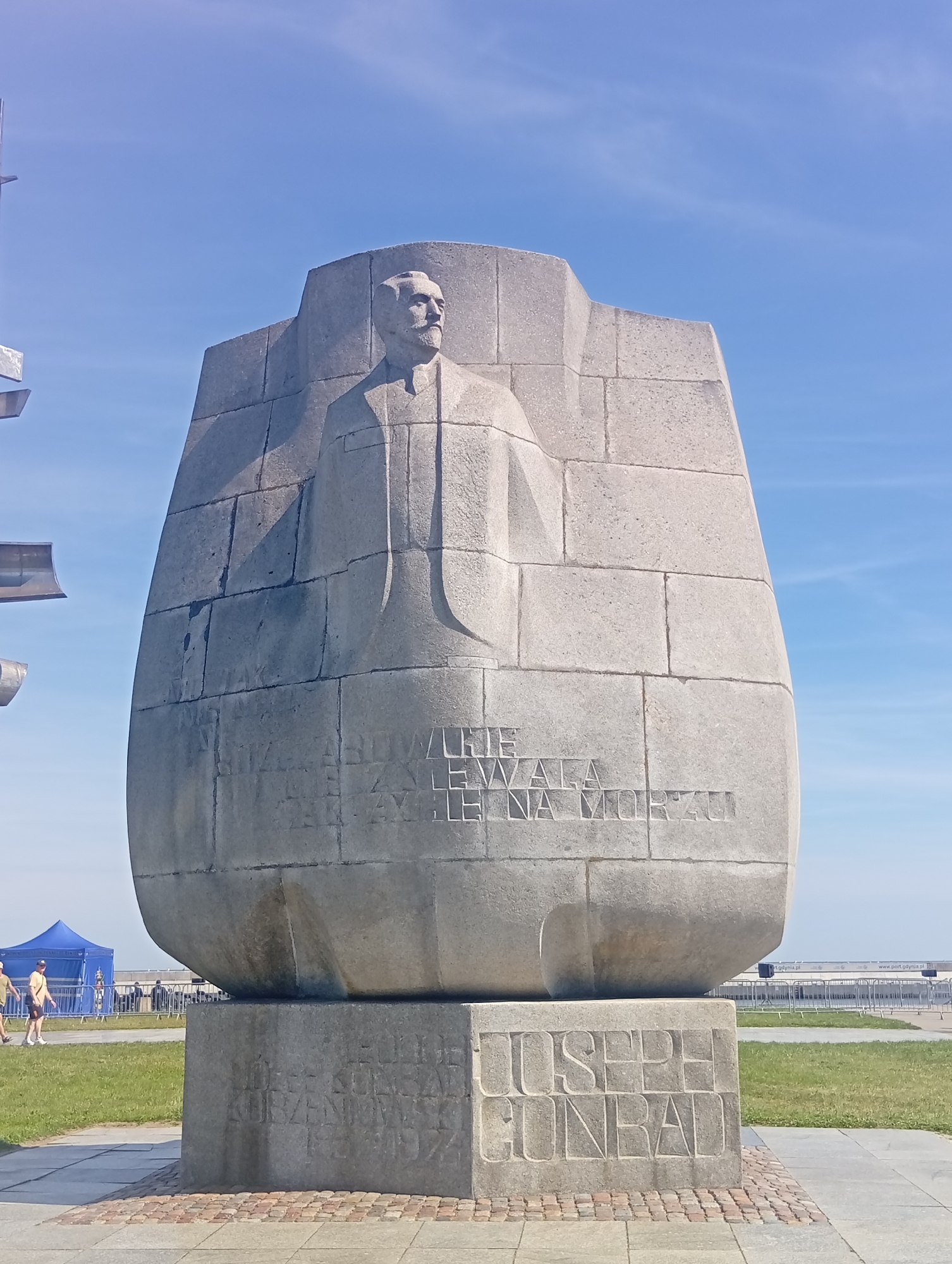 The image shows a large, weathered stone monument in Gdynia, Poland, depicting a bust of a man with a mustache, dressed in a suit and tie. The monument is set against a clear blue sky. The stone is carved to resemble blocks, and inscriptions are visible on its surface. Below the bust, an inscription reads "Józef Konrad". To the right of this name, another inscription partially reads "CONRAD". The base of the monument also has carved text, though it is partially obscured and weathered.
In the background, there is a grassy area, a paved walkway, and in the far distance, a blue tent and a fence with people visible behind it. The overall setting appears to be an outdoor public space, possibly a waterfront area given the industrial structures in the far background.