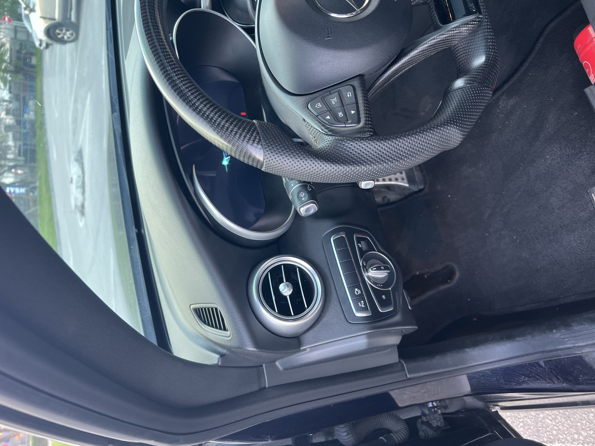The image shows the interior of a car, specifically the dashboard, steering wheel, and part of the door. The steering wheel is black with carbon fiber accents. The dashboard features an air vent and several control buttons. The environment visible through the windshield appears to be an urban setting. There is no indication of this being in Plovdiv, Bulgaria. There are no people visible in the image.