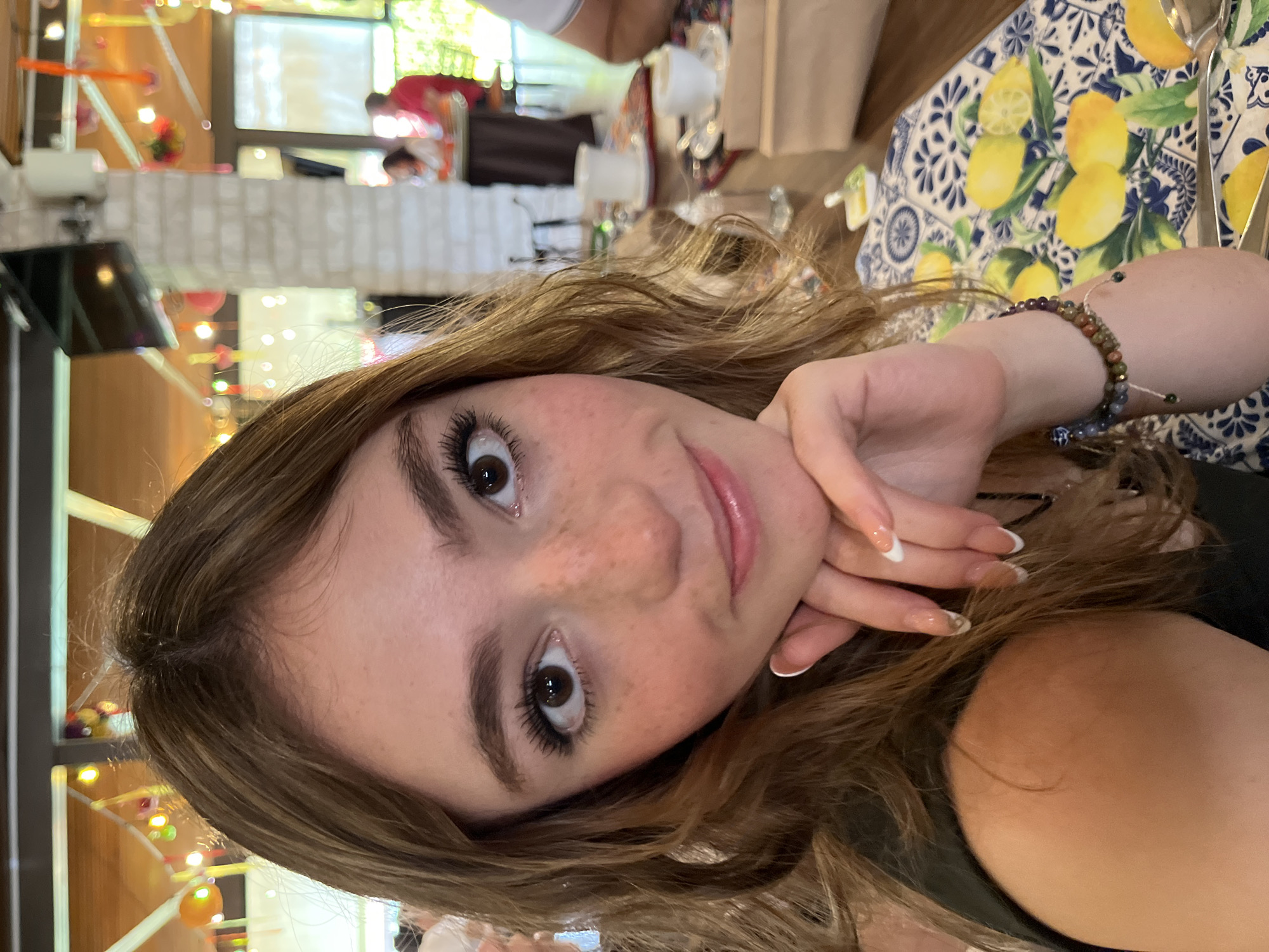 A young woman with light brown hair and freckles poses for a selfie at a restaurant. She is leaning her head on her hand, revealing a bracelet. The background shows other patrons, tables with floral patterned cloths, and outdoor light coming through windows. The location appears to be a restaurant in Unknown, Unknown.