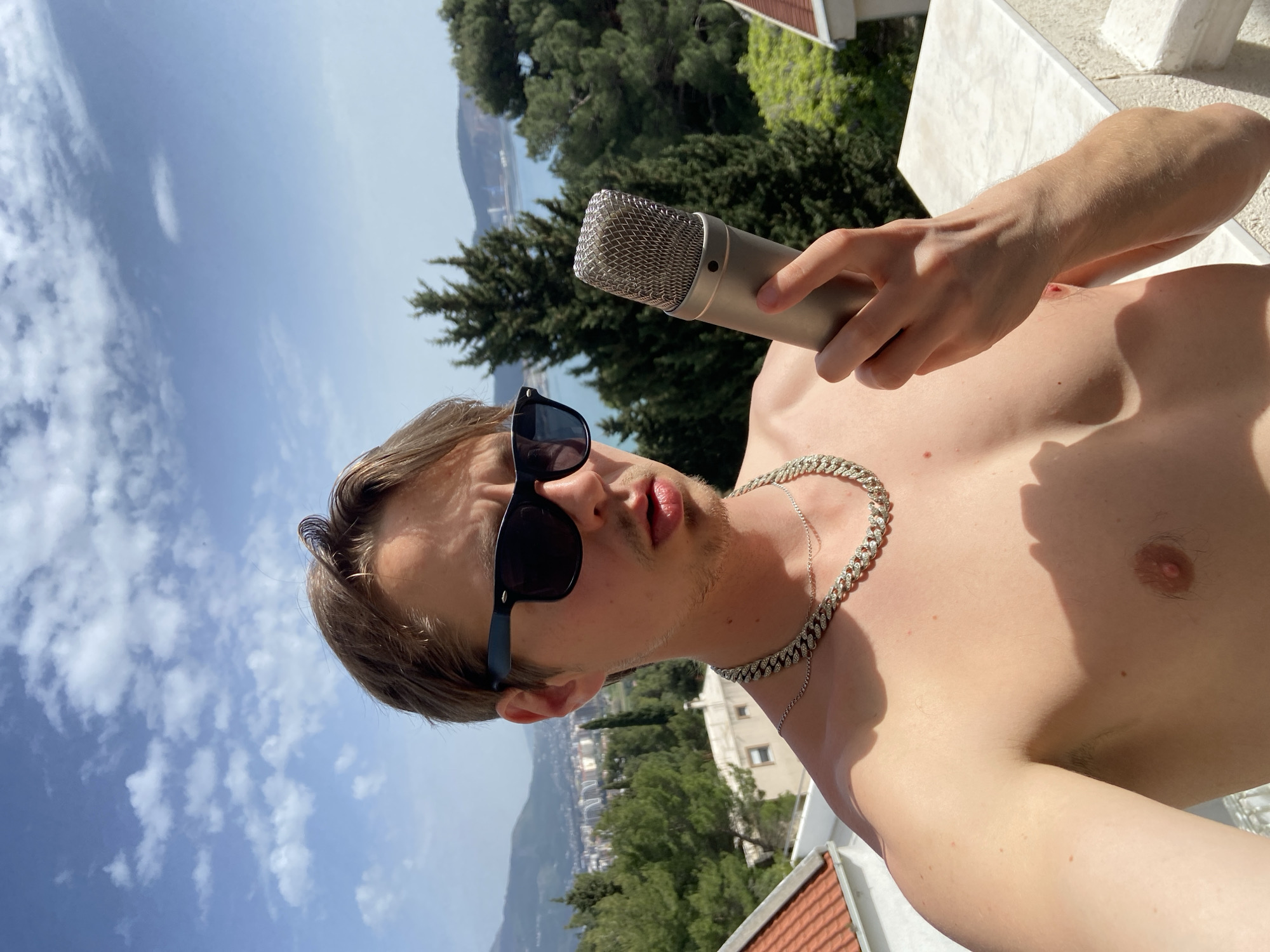 A shirtless man with sunglasses and a silver chain holds a microphone in his right hand. The background includes a cityscape with trees, mountains, and a body of water. The location is likely Šušanj, Montenegro, based on the landscape and architecture.