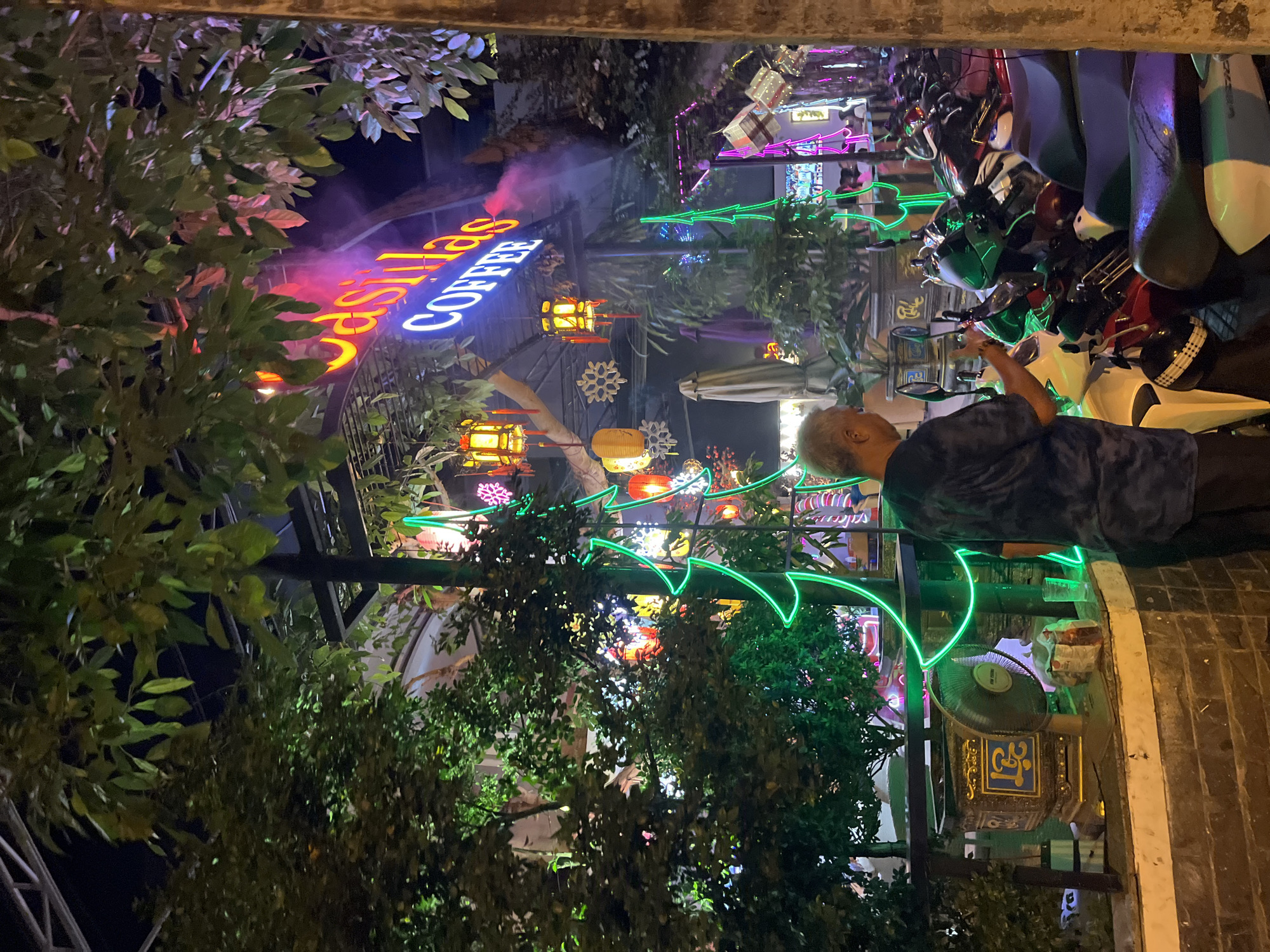 This is a night scene outside "J'sillas Coffee" in Nha Trang, Vietnam. A man with gray hair, wearing a dark blue t-shirt, stands on the left, facing a row of parked motorcycles. He appears to be looking at something in his hands, possibly a phone. In front of him, a stone planter box with ornate gold lettering and a fan are visible. Neon green lights outline a Christmas tree shape to his right. The coffee shop's sign, illuminated in neon pink and blue, is visible in the background. Several paper lanterns hang from the shop's awning. The area is decorated with string lights and other colorful illuminated elements.